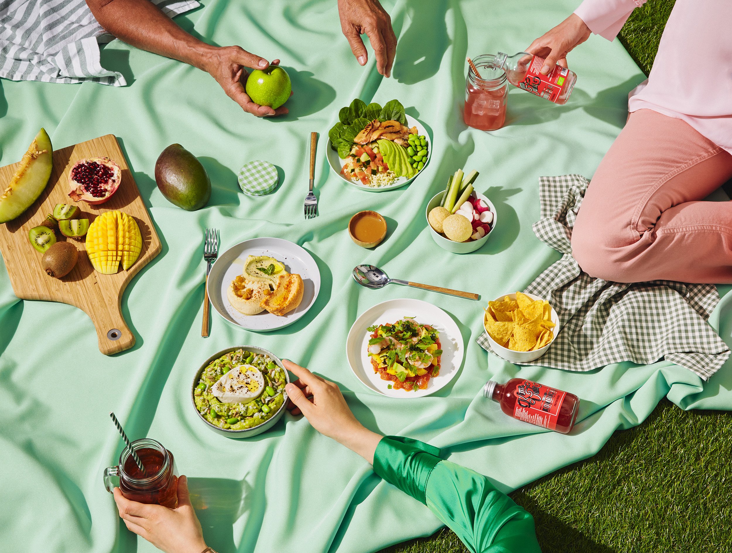 Mesa de picnic con una variedad de comida y bebidas, incluyendo frutas, ensaladas, chips, y jugos, en una manta verde en un día soleado.