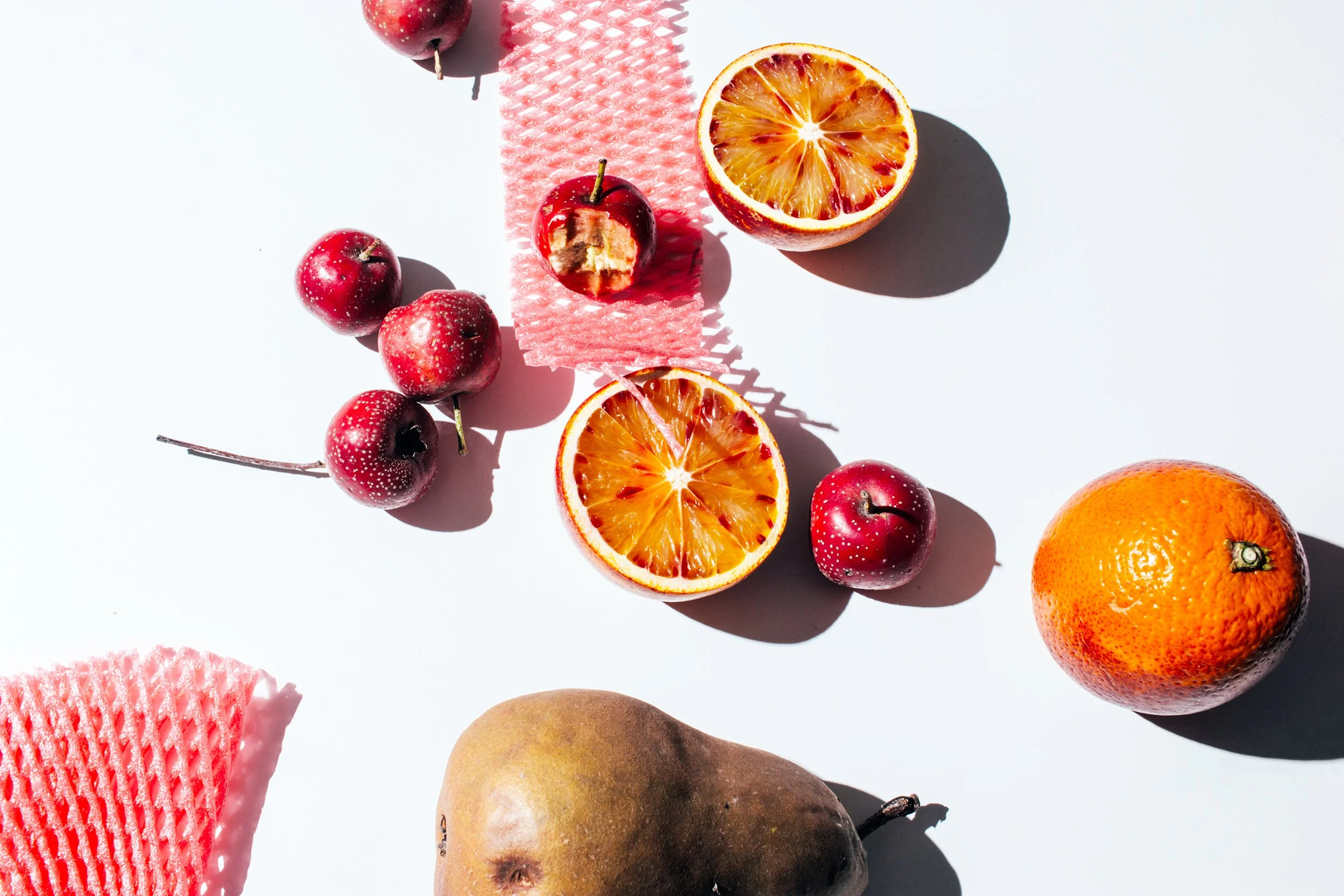 Variedad de frutas en una superficie blanca, incluyendo manzanas rojas, naranjas cortadas, una pera y un melón, además de una gasa rosa y su sombra.