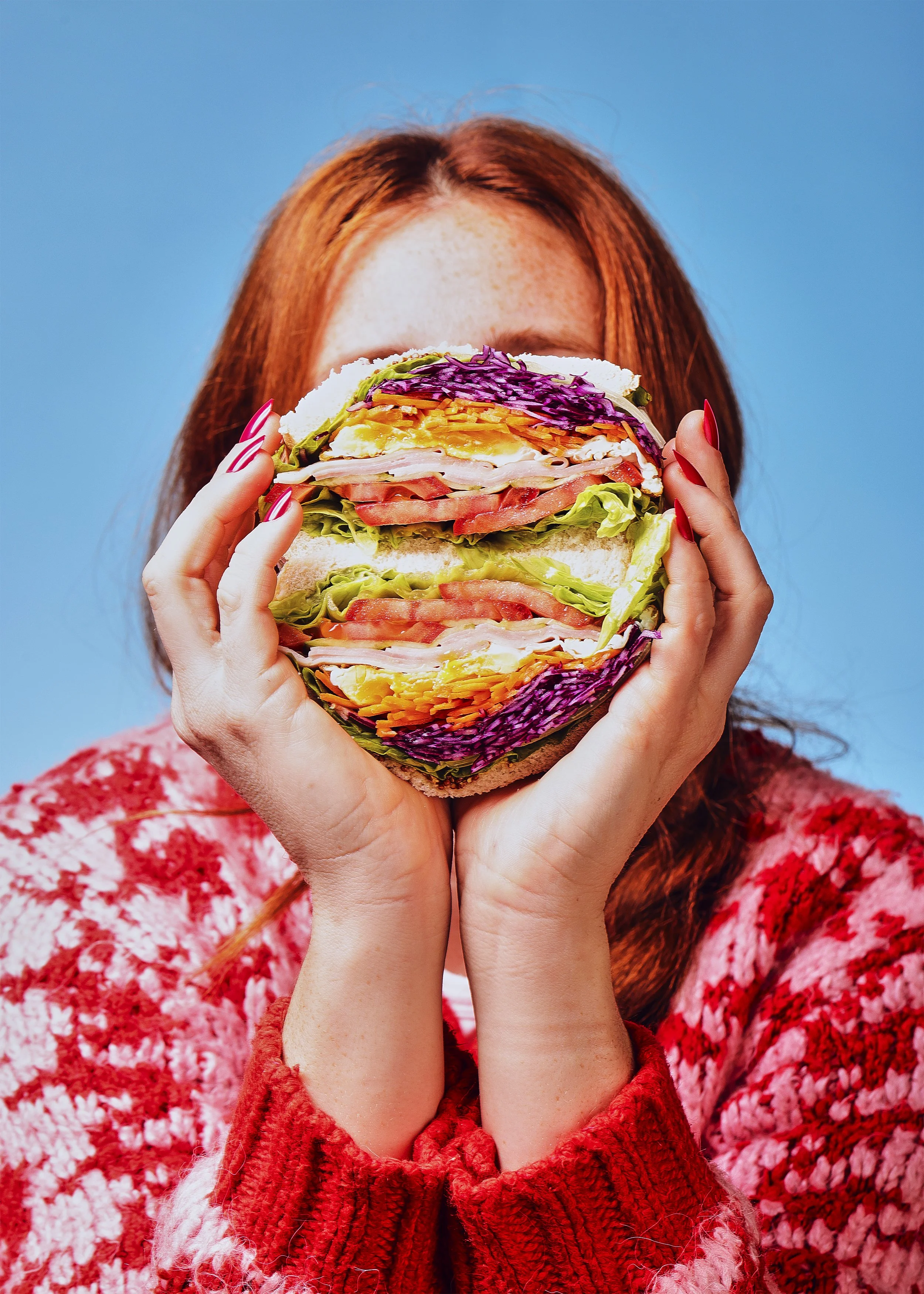 Una mujer con cabello rojo sostiene un sándwich gigante y colorido frente a su rostro, con un fondo de cielo azul.