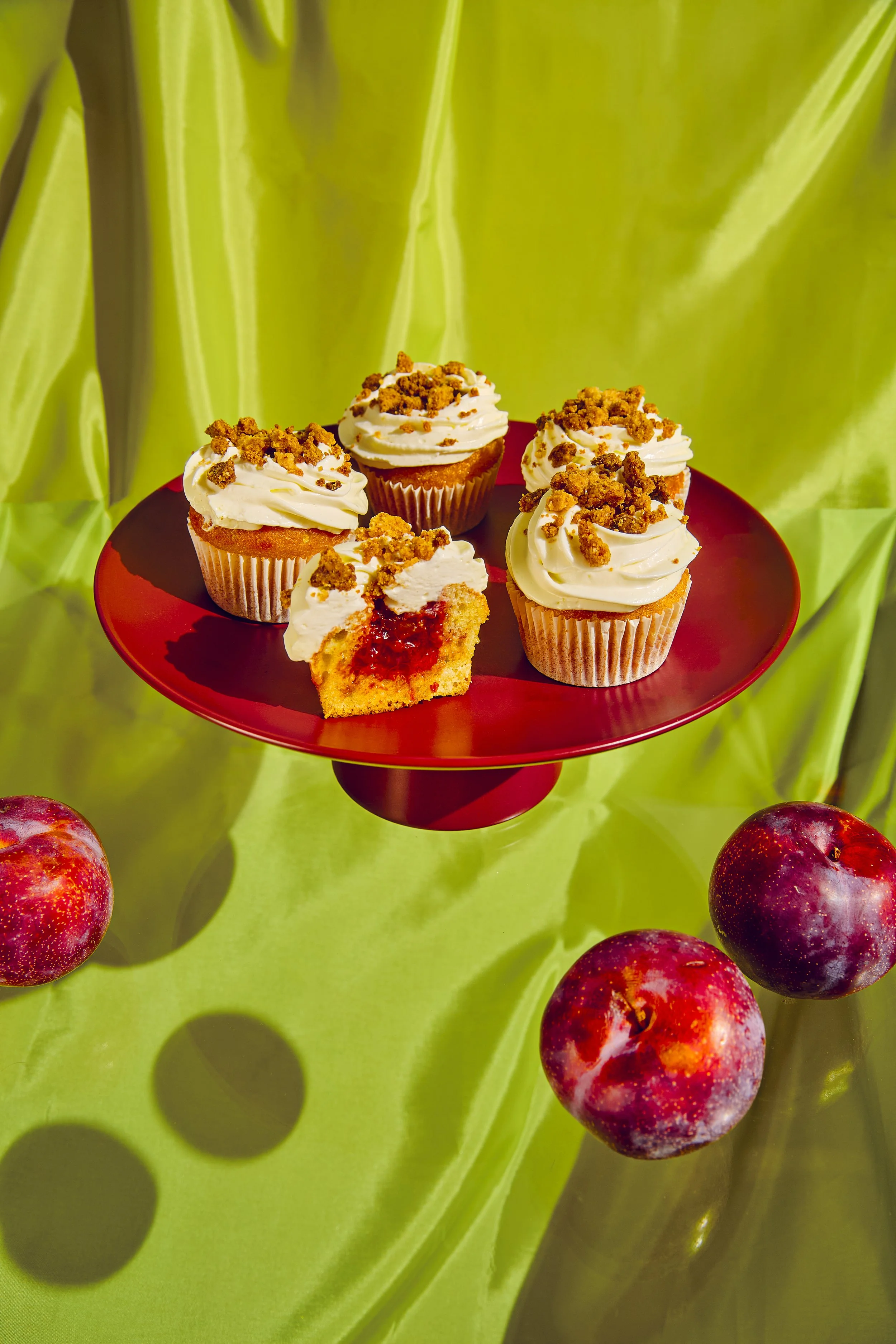 Cuatro cupcakes con glaseado y crumble en un plato rojo, con una rebanada mostrando relleno de mermelada de cereza, en un fondo de tela verde con cerezos.