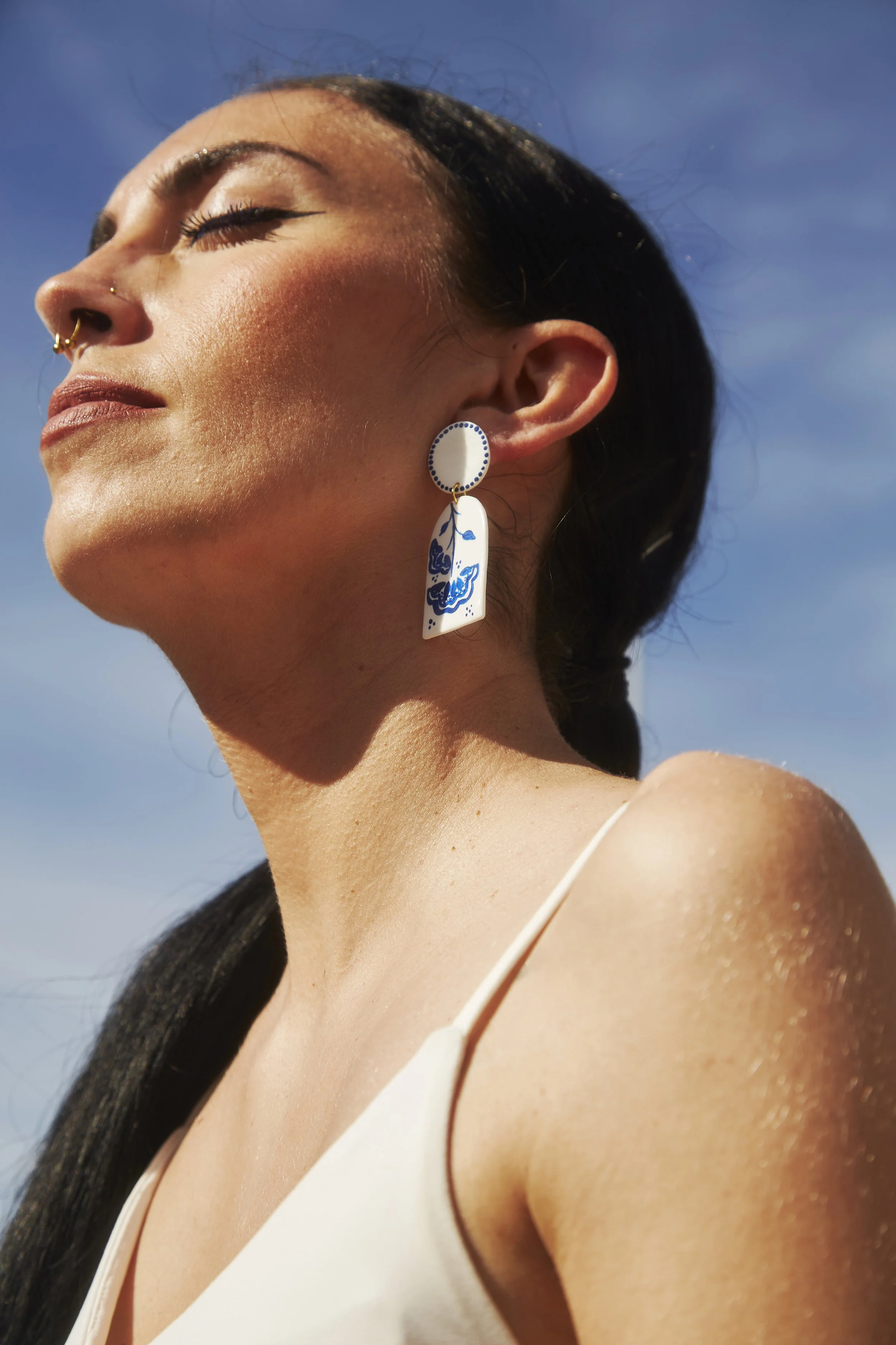 Mujer con piel bronceada y cabello oscuro, usando pendientes grandes y brillando con los ojos cerrados bajo el sol.