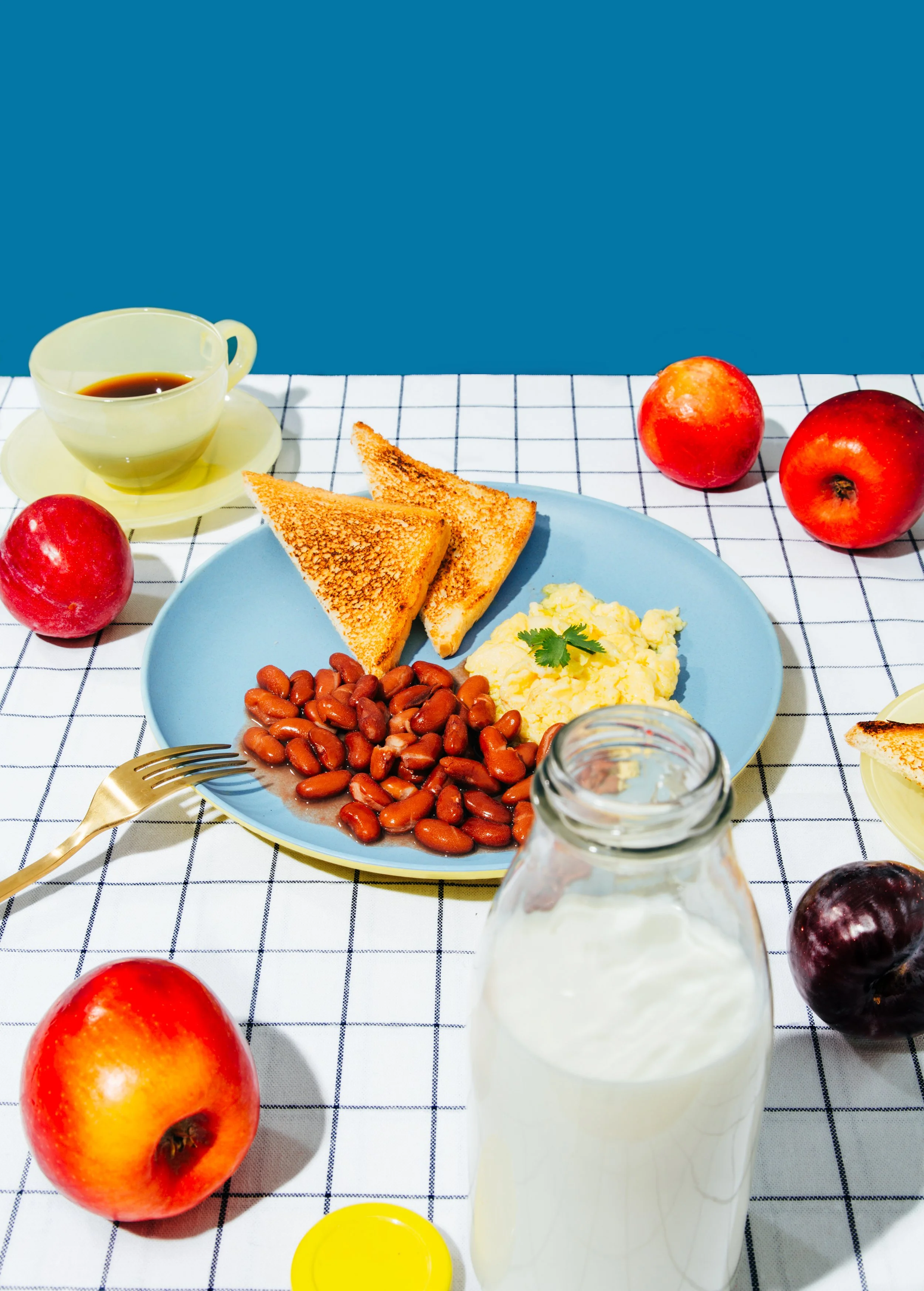 Desayuno con huevos revueltos, frijoles, tostadas, manzanas, leche, café y jarra de leche en una mesa con mantel de cuadros blancos y azules.