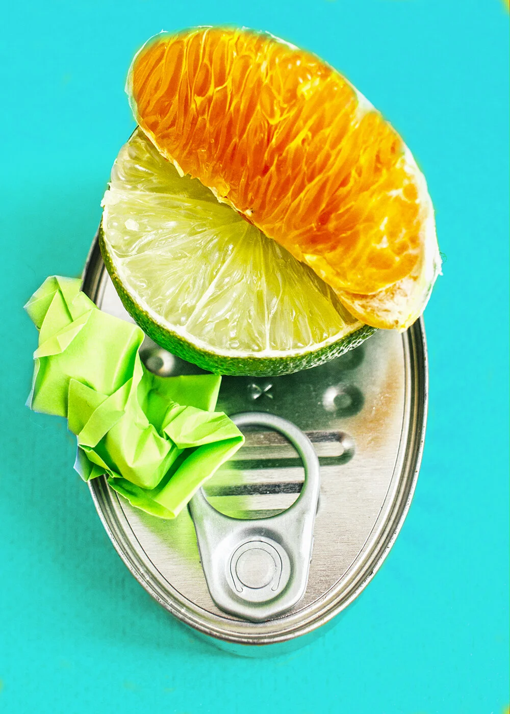 Trozo de lima con un gajo de mandarina encima, y un trozo de papel de color verde al lado, todo sobre una lata de aluminio