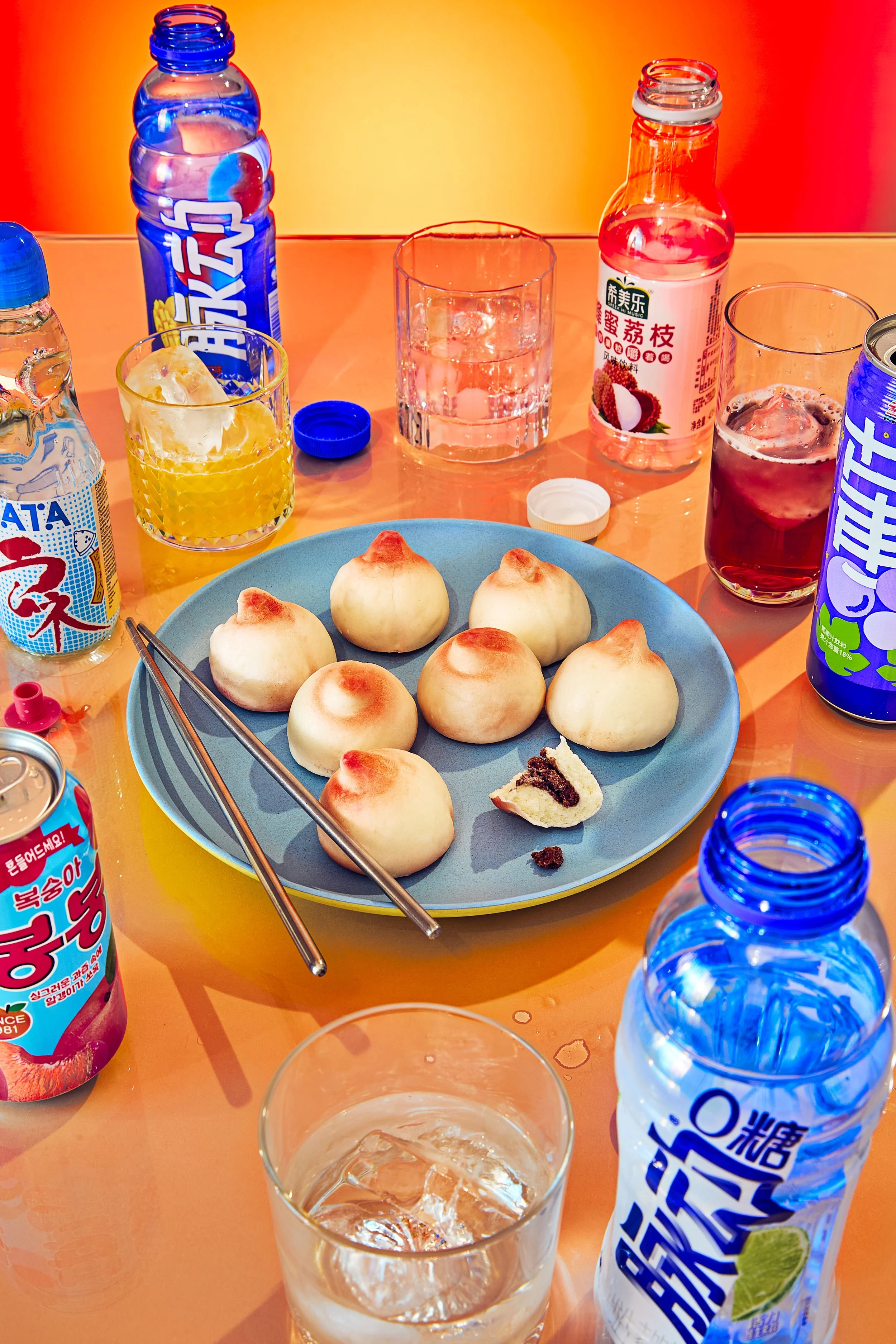 Plato de dumplings chinos y refrescos en botellas de diferentes sabores y colores, algunos vasos con hielo y líquido, y cucharas y palillos de metal en un ambiente colorido.