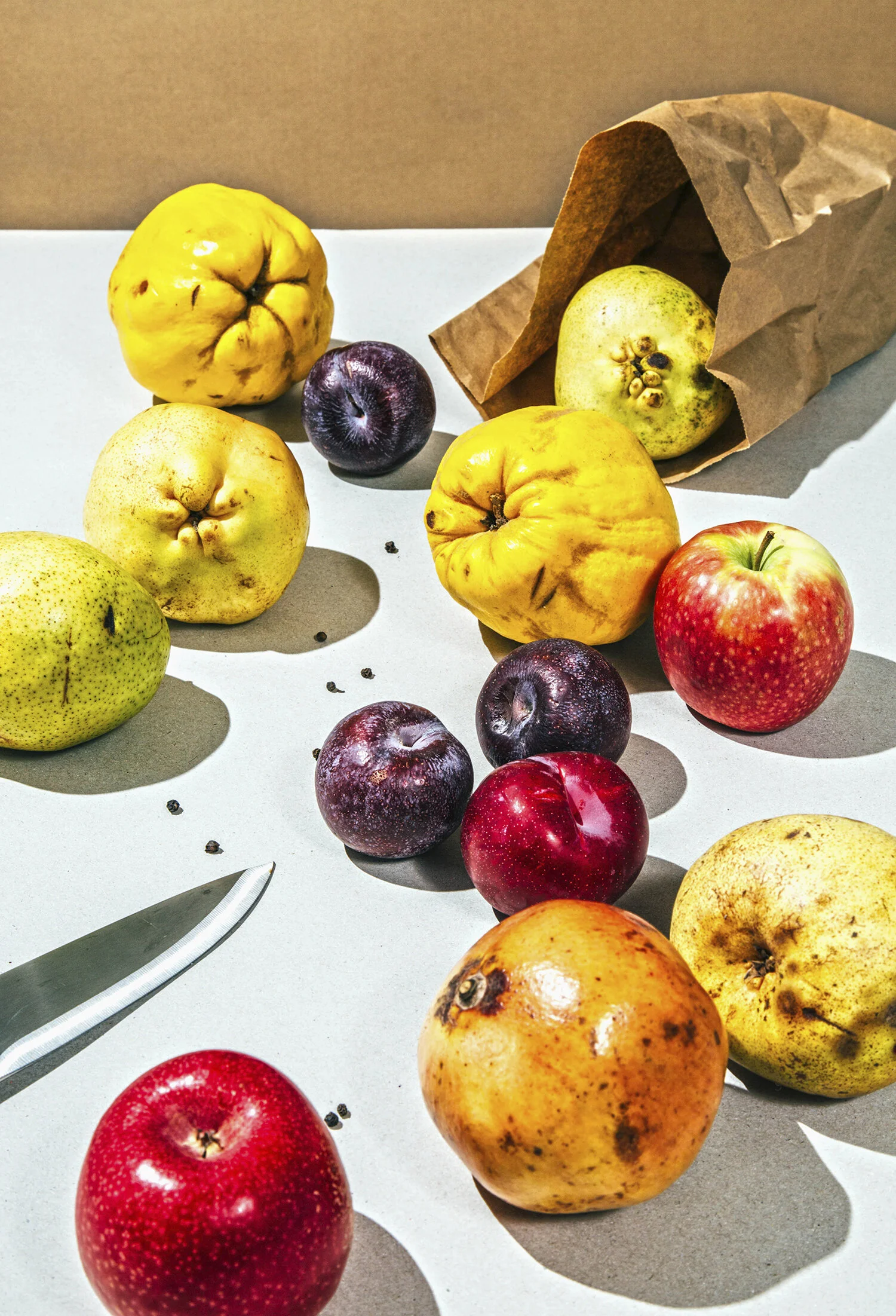 Varias frutas en una mesa, incluyendo manzanas, peras, uvas y algún fruto de color amarillo, con sombra y en un papel de supermercado, en un ambiente con luz natural.