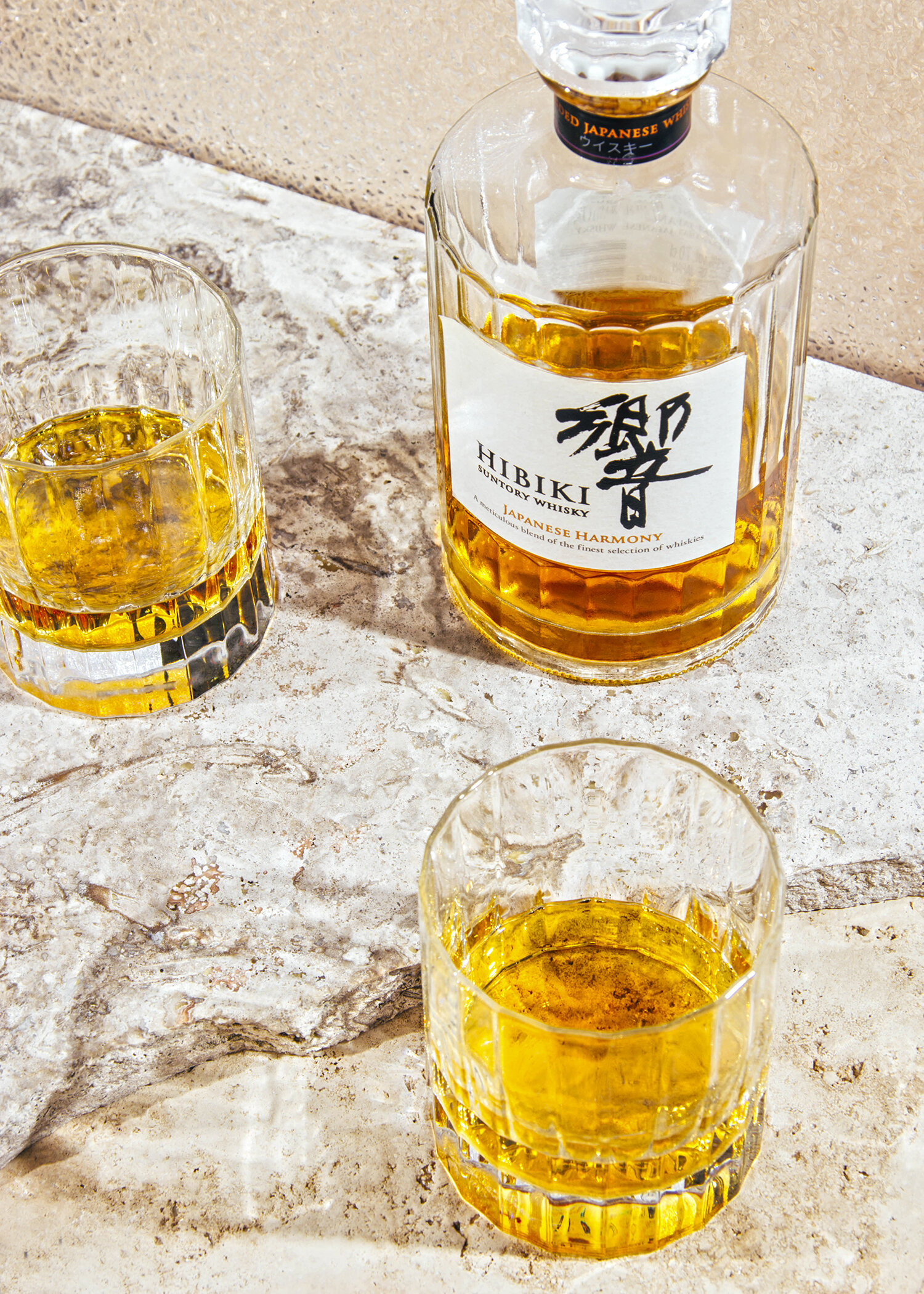 Botella y vaso de whisky Hibiki Suntory con etiqueta en japonés, en una superficie de piedra clara.