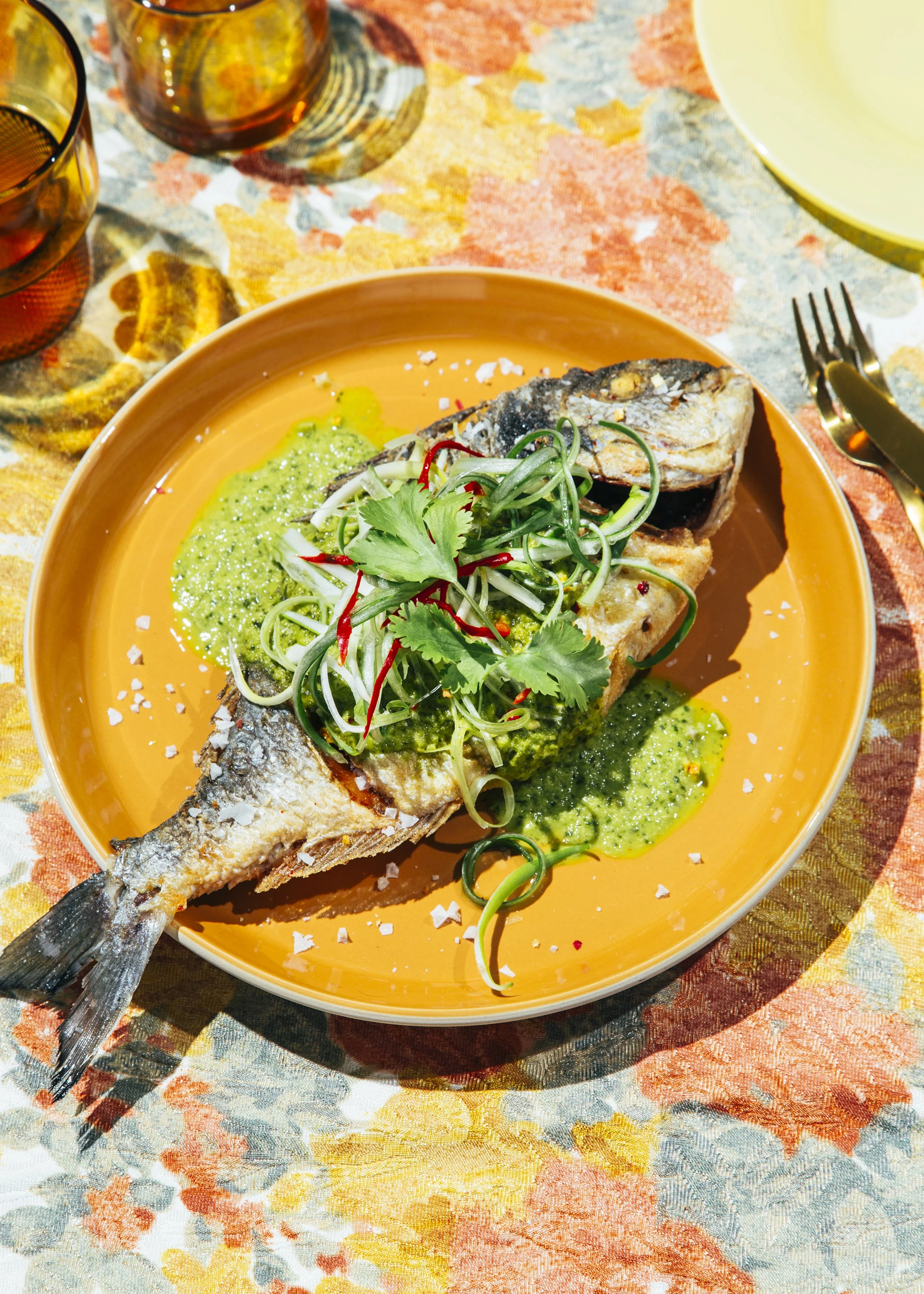 Pescado frito servido con salsa de aguacate, cilantro, cebolla y chiles en un plato naranja sobre una mesa con manteles coloridos.