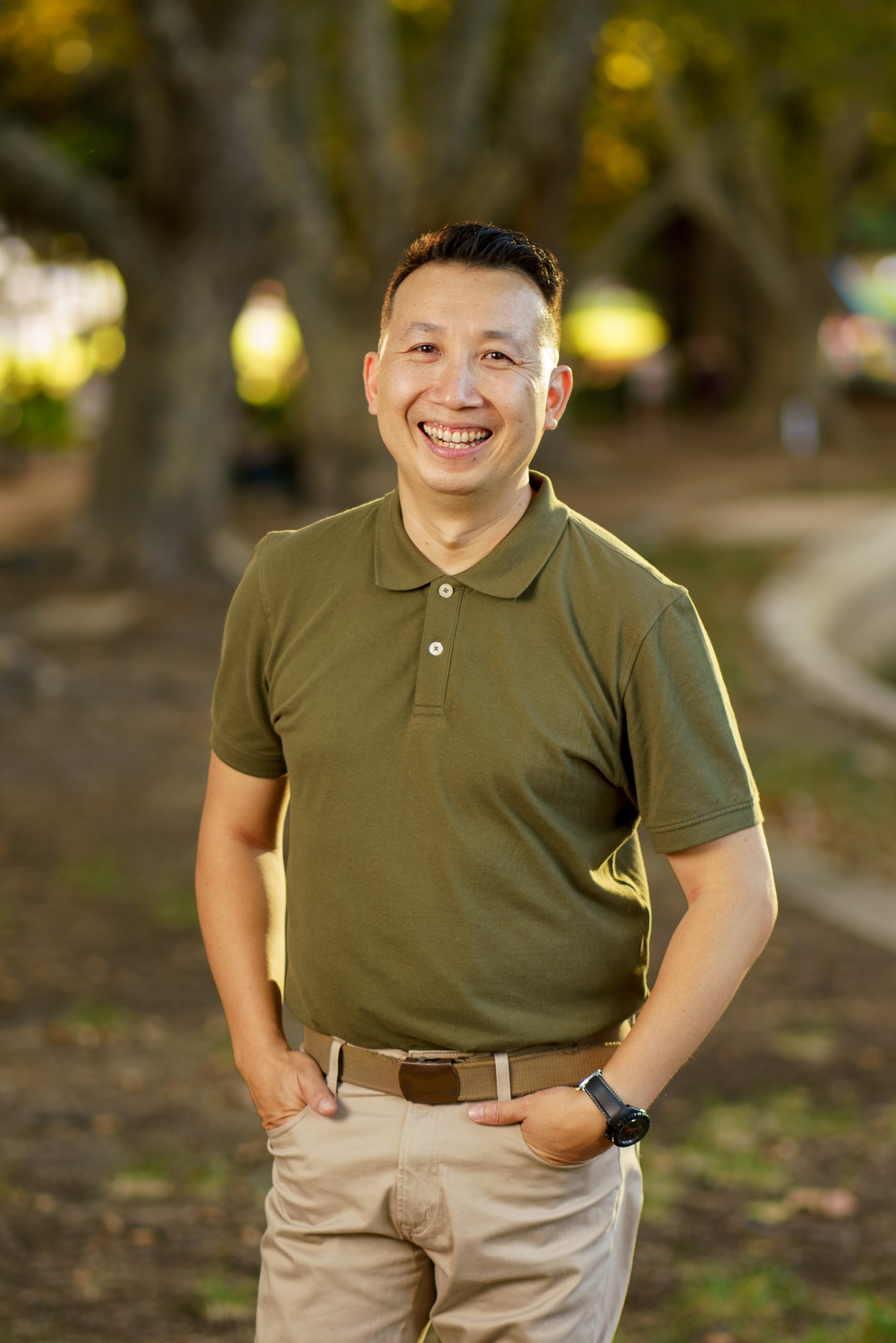 A smiling man with short black hair, wearing an olive green polo shirt and khaki pants, standing outdoors in a park with trees in the background.