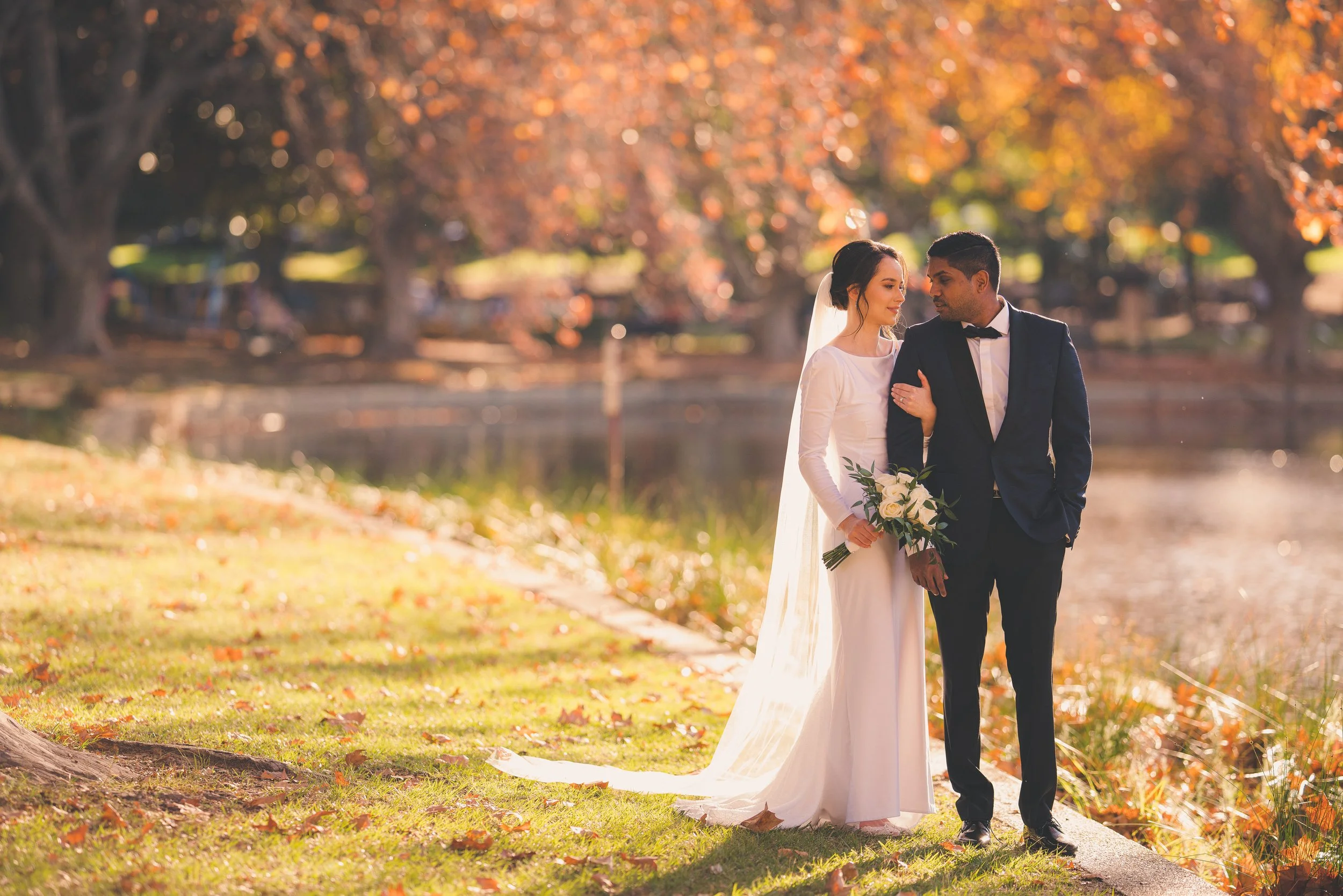 A bride and groom walking arm in arm along a lakeside path during autumn, surrounded by fall foliage with orange and yellow leaves.