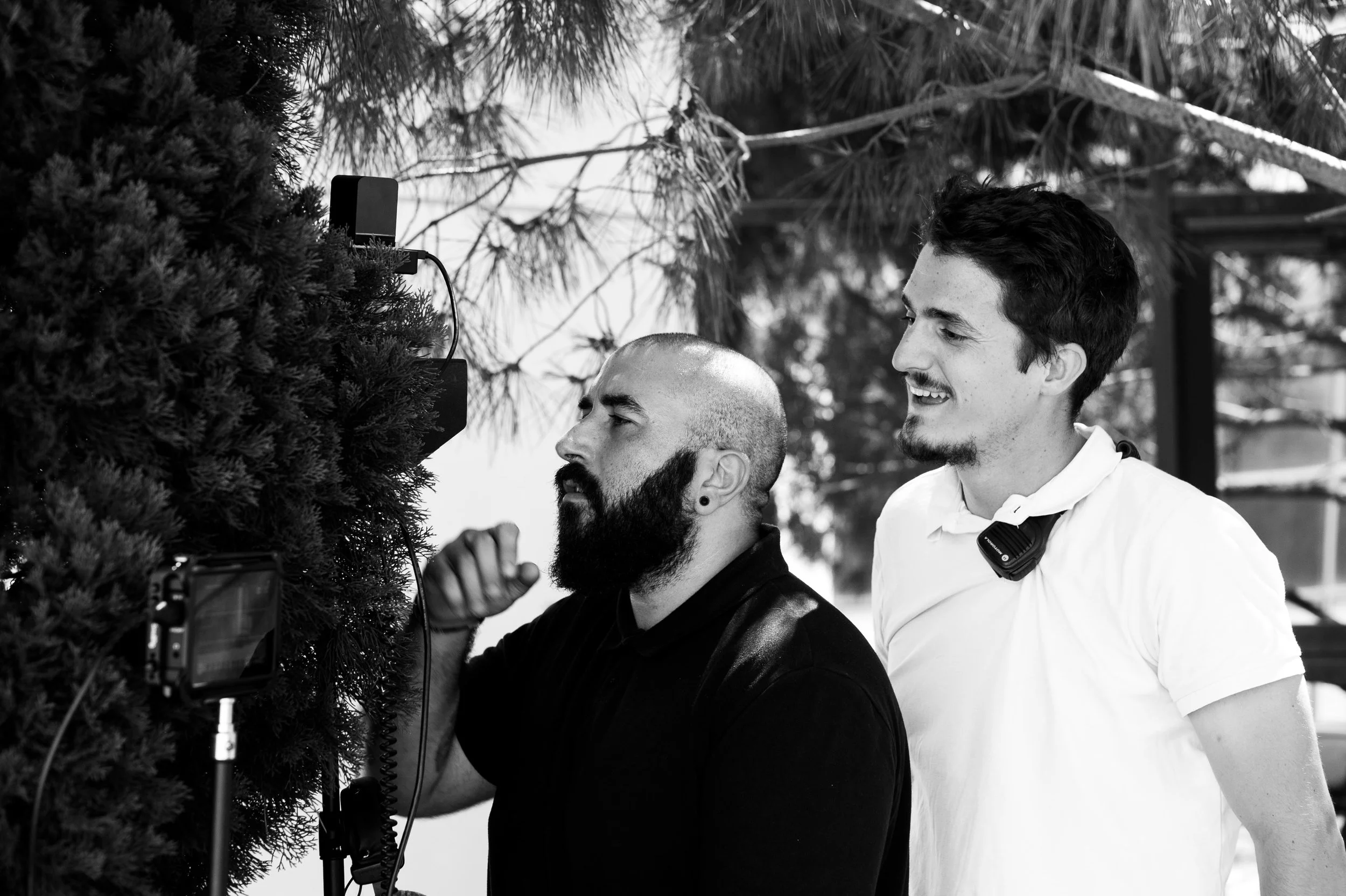 Deux hommes en extérieur, un vérifiant un appareil photo ou une caméra, parmi des arbres, en noir et blanc.