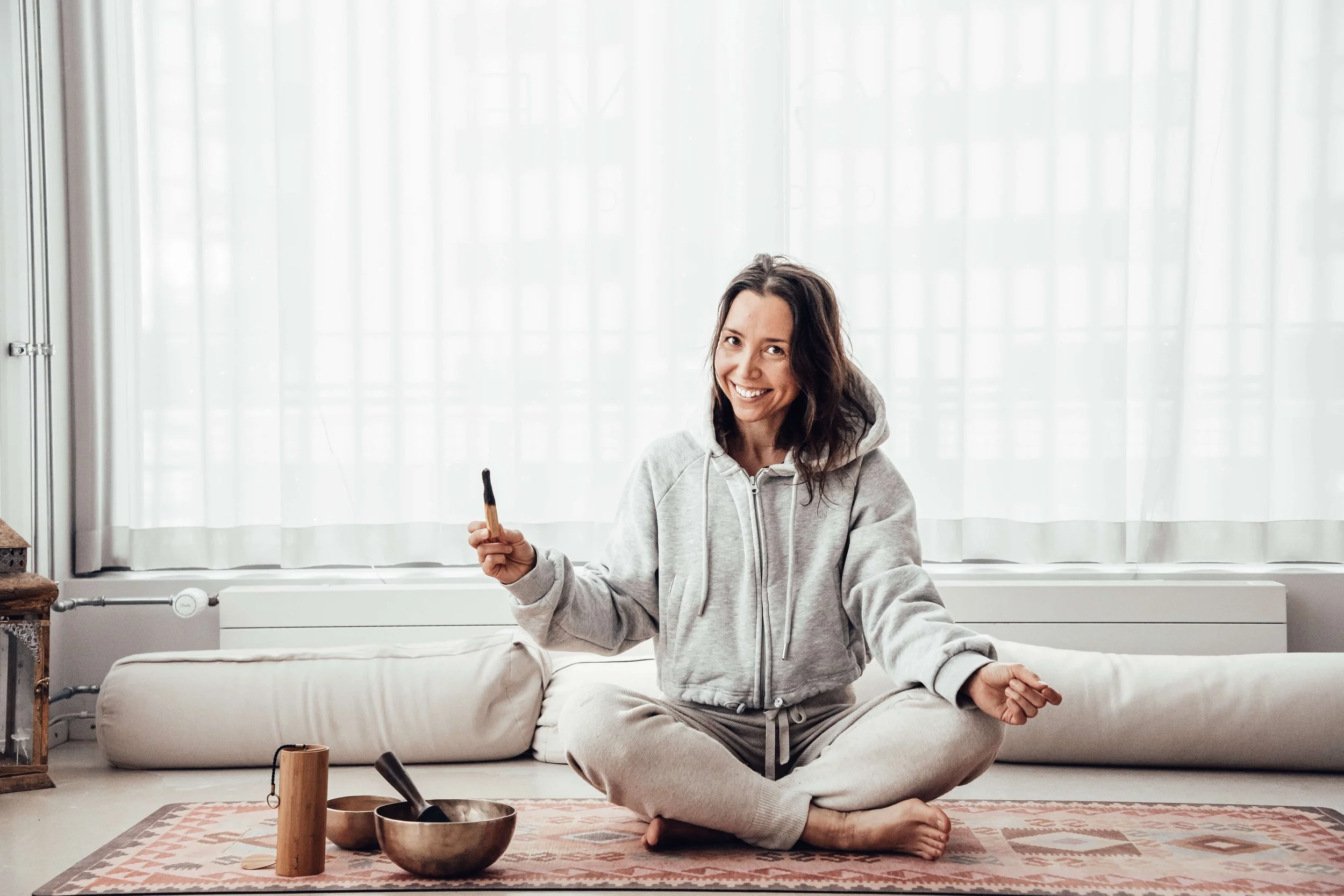 Joogaohjaaja istuu lattialla meditaatioasennossa, hymyilee, taustalla suuri ikkuna ja valkoinen verho. Naisen vieressä äänimaljoja