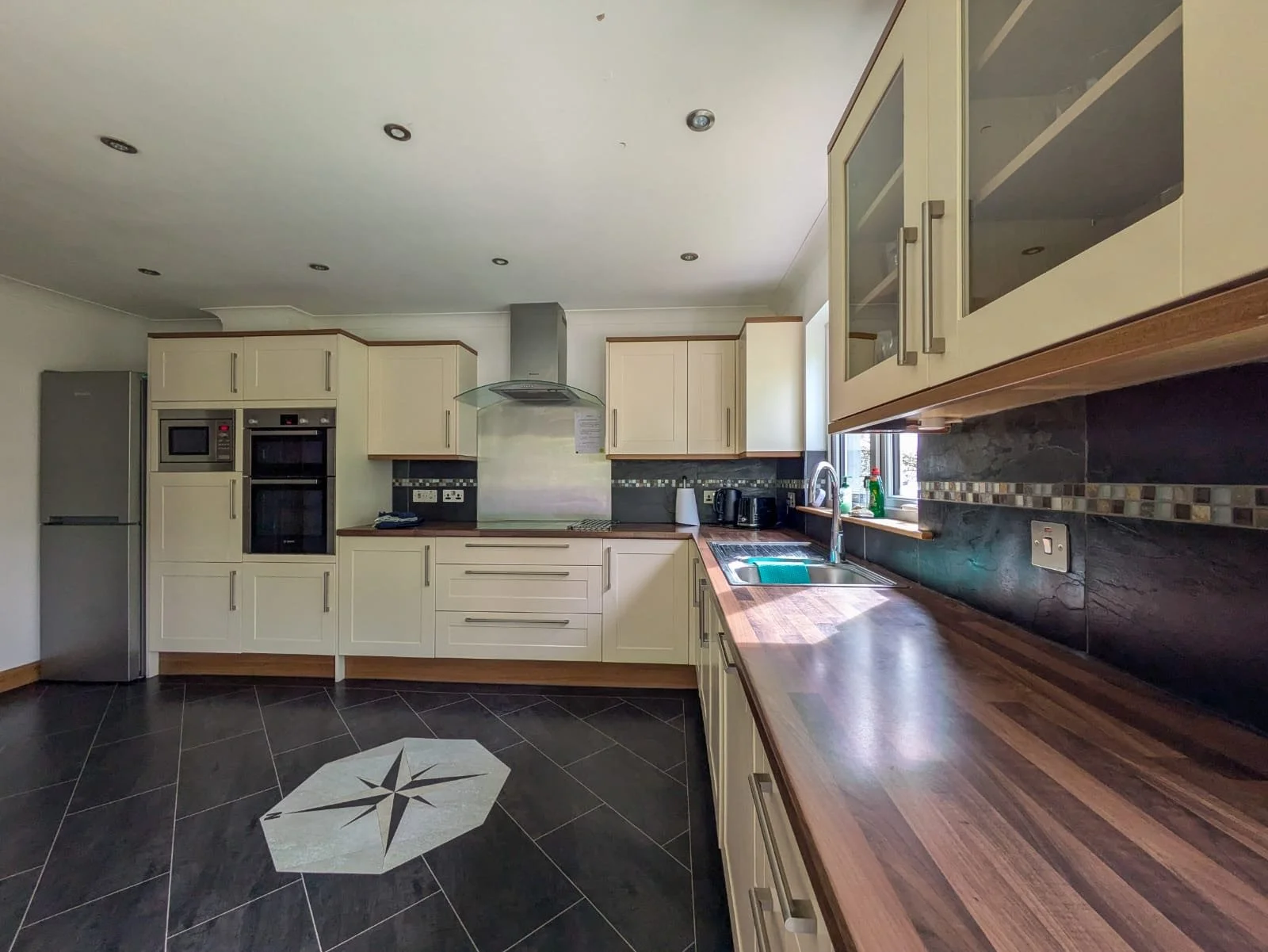 A modern kitchen with cream-colored cabinets, a wooden countertop, black backsplash, and built-in appliances including a microwave and oven. There is a stainless steel refrigerator, a range hood over the stove, a sink under a window, and black floor 