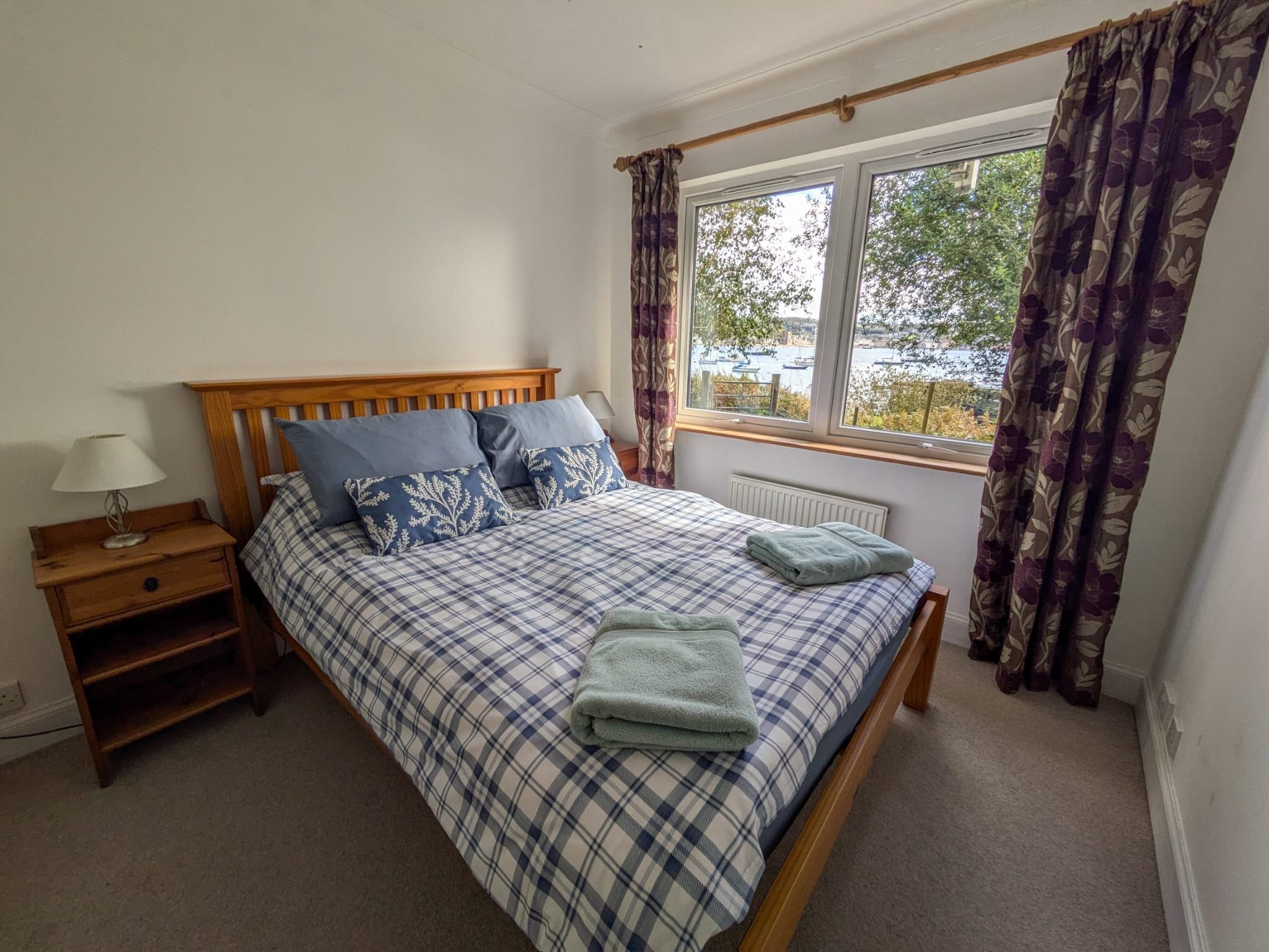 A bedroom with a wooden bed frame, blue and white plaid bedding, and four pillows with blue covers. There are two folded towels on the bed. To the left, a wooden nightstand with a small lamp. The room has a large window with purple floral curtains, s