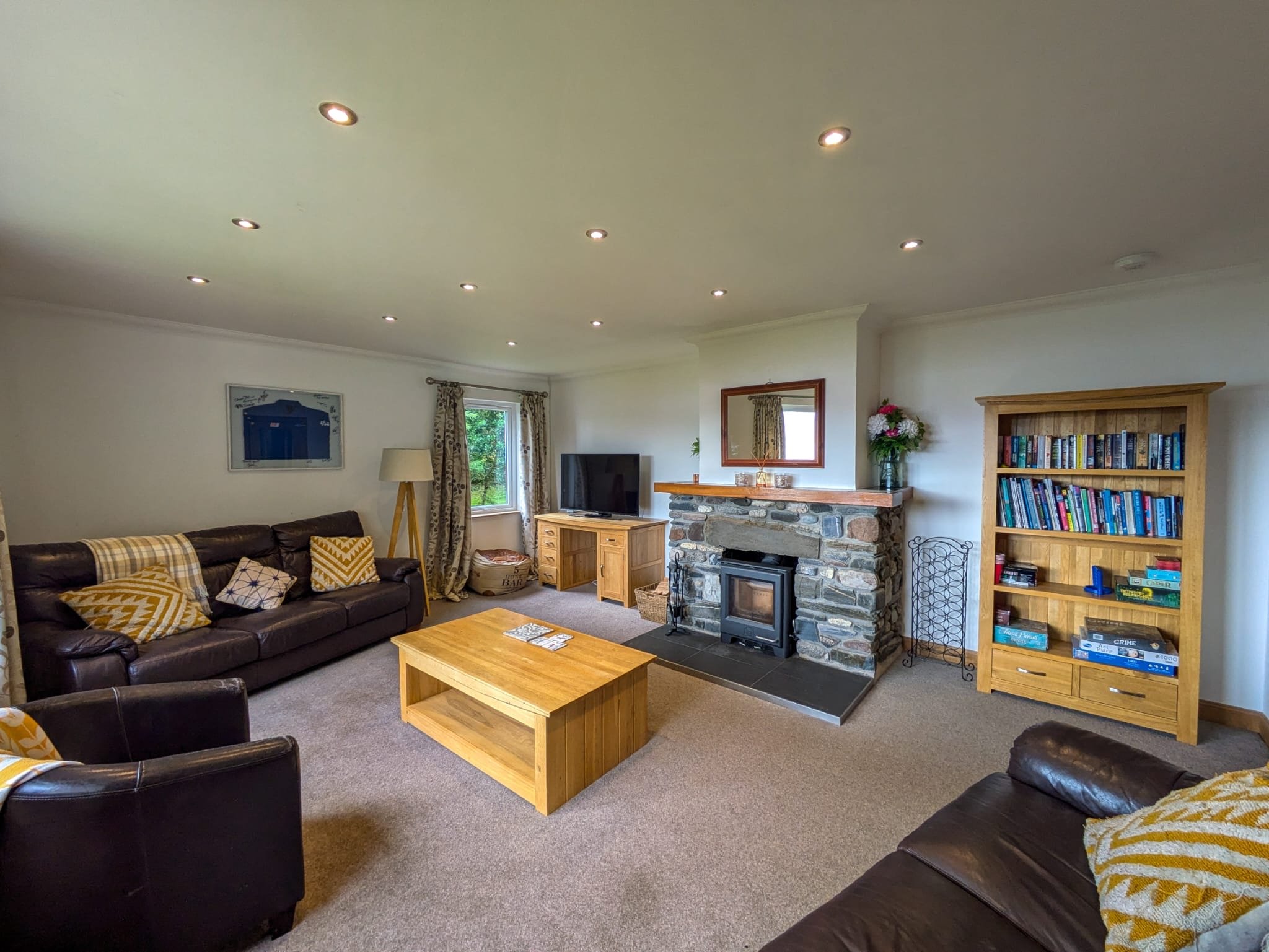 A cozy living room with a leather sofa, a wooden coffee table, a stone fireplace, a bookshelf, a flat-screen TV, and patterned curtains.