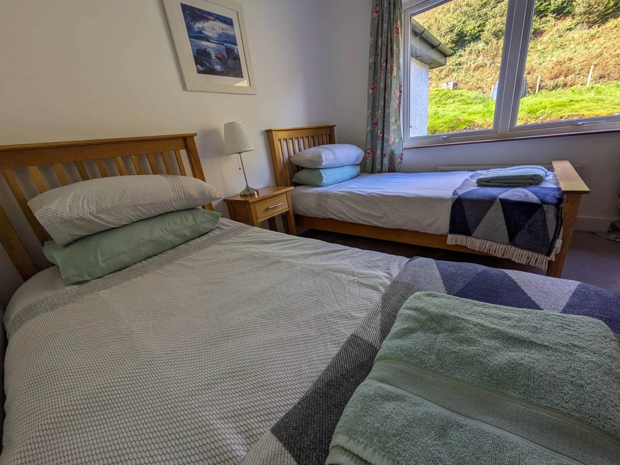 A cozy bedroom with two wooden beds, a nightstand with a lamp, a picture on the wall, and a large window showing a landscape of green grass and trees.