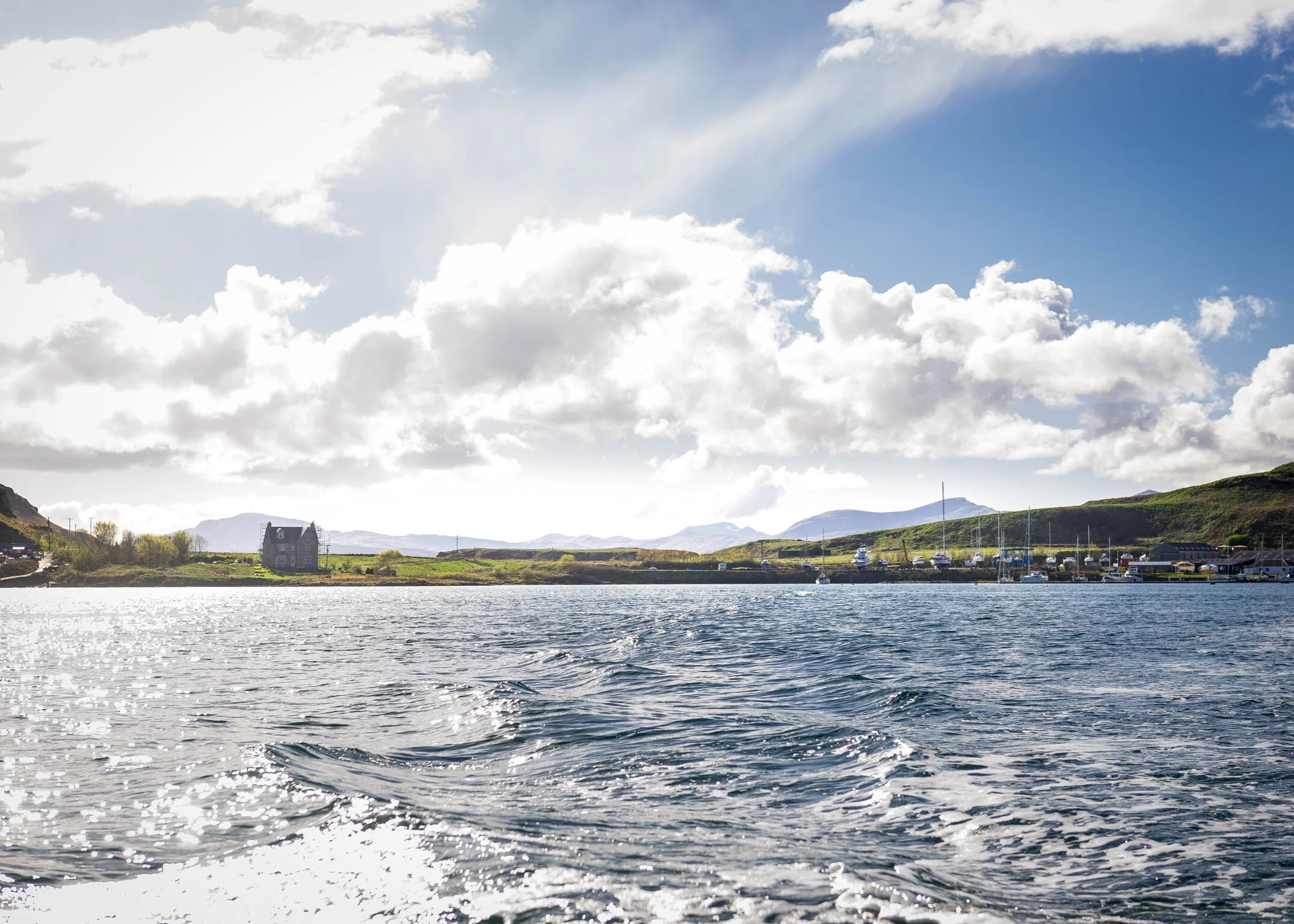 A scenic view of a body of water with gentle ripples, a distant shoreline with a house, green hills, mountains in the background, and a partly cloudy sky.