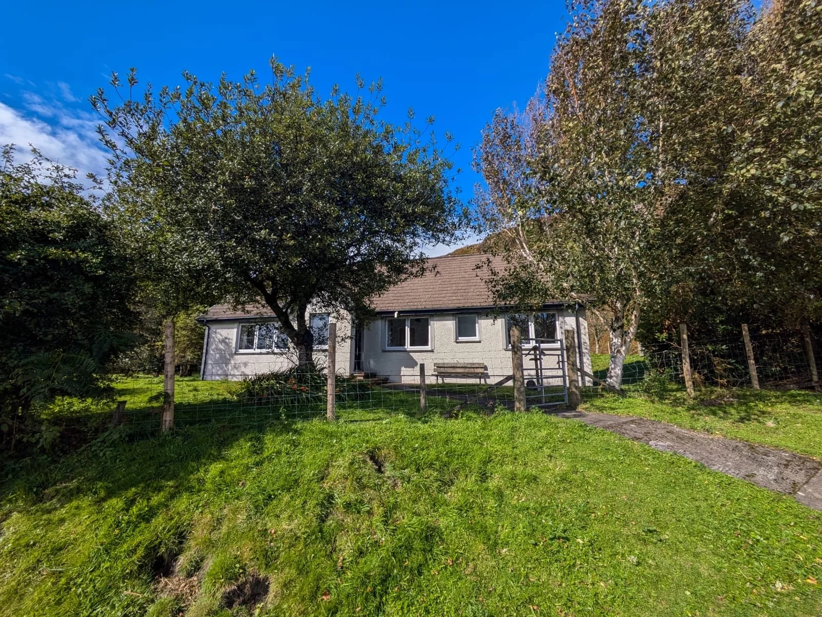 A single-story house with a brown roof, white exterior walls, and multiple windows, surrounded by green grass, trees, and a wooden fence under a clear blue sky.