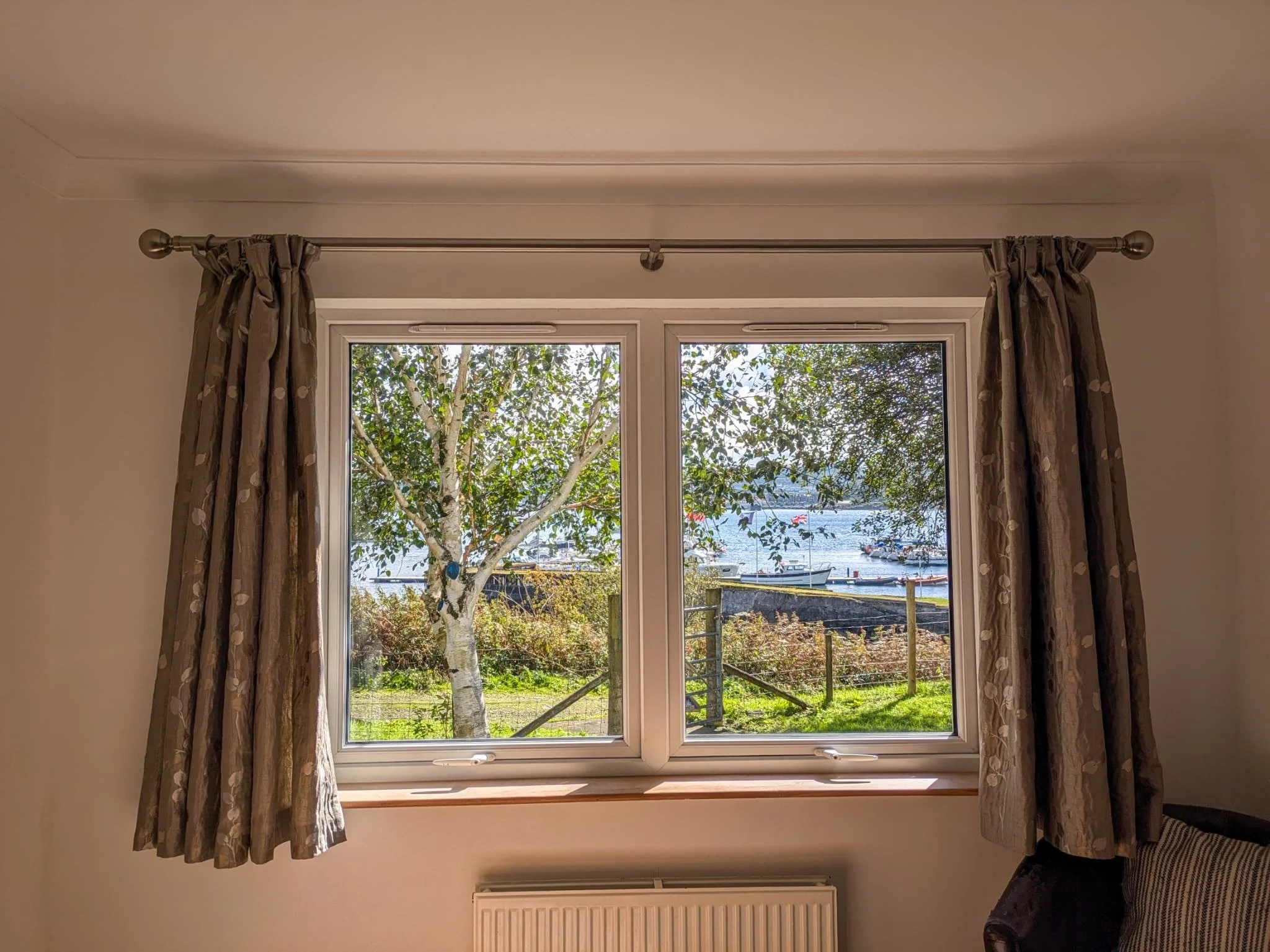 A window with beige curtains showing a view of a tree, a body of water, boats, and a fence outside.