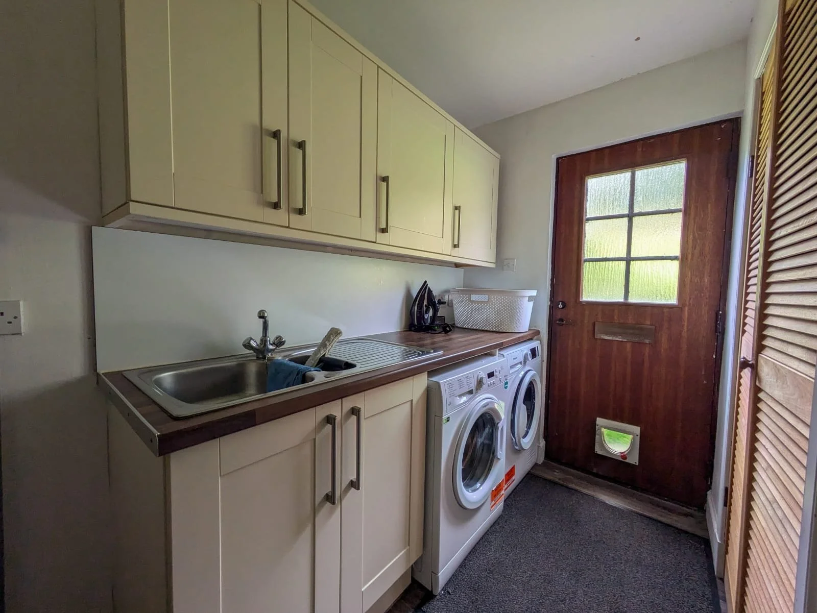 Laundry room with cream upper cabinets, a stainless steel sink, a washing machine, a wooden door with a window, and a louvered closet door.