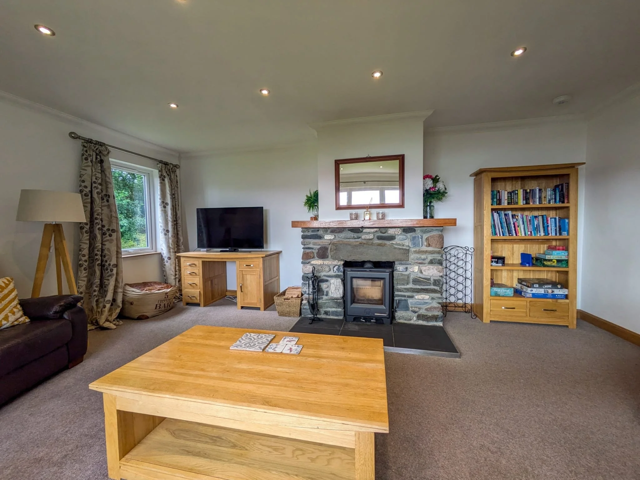 Living room with stone fireplace, wooden furniture, TV, bookshelf, window with floral curtains, and carpeted floor.