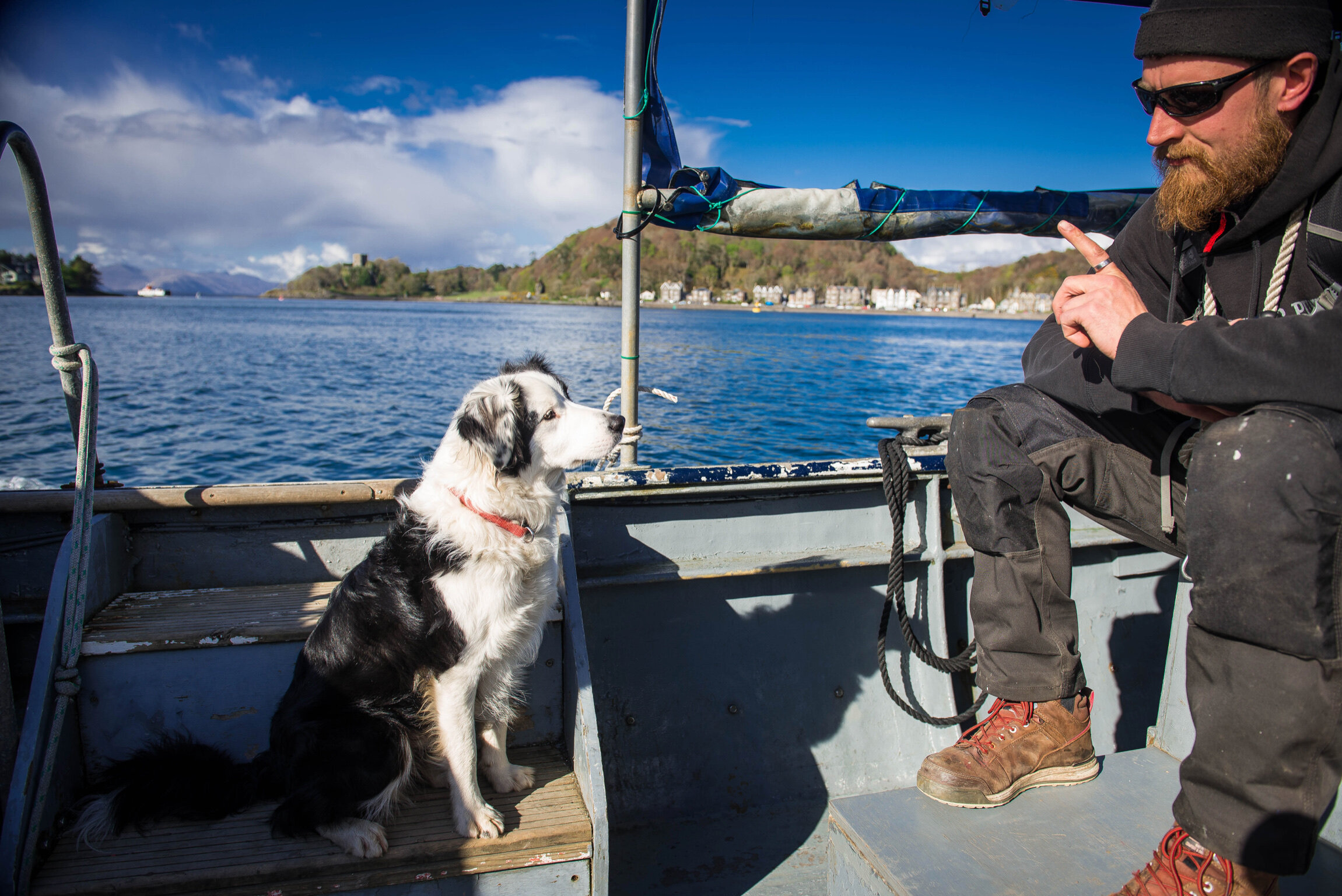 A man with a beard, wearing sunglasses and a black hoodie, sitting on a boat with a black and white dog sitting next to him. The boat is on a body of water with a scenic backdrop of hills and buildings under a partly cloudy sky.