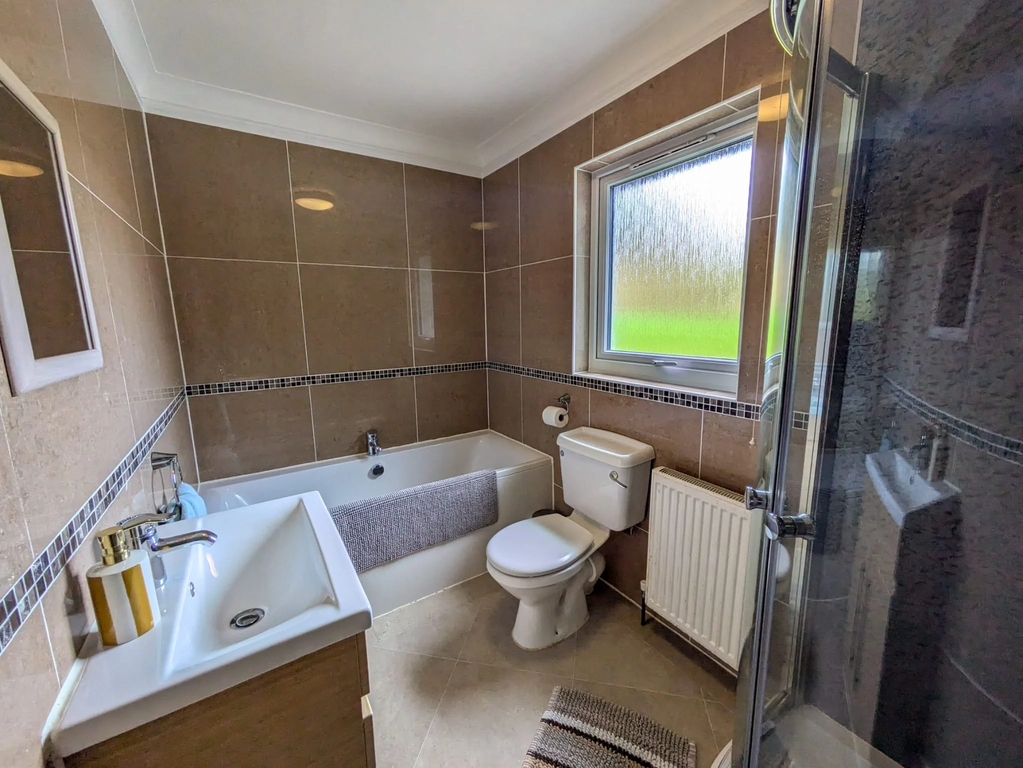 Bathroom with brown tiled walls, toilet, bathtub, small sink, and a window with frosted glass.