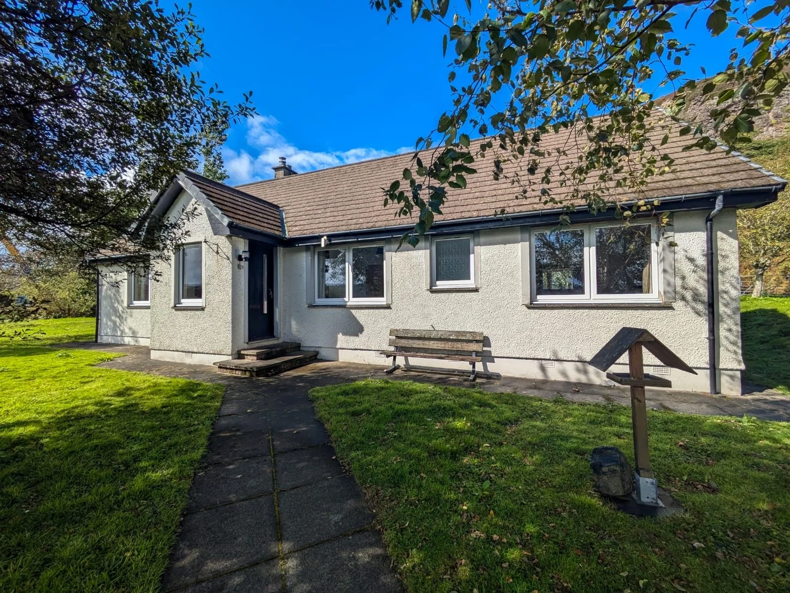 A single-story house with cream-colored stucco walls, a brown shingled roof, and multiple rectangular windows. There is a concrete sidewalk leading to the front door, a wooden bench, and a small wooden signpost in the yard. The house is surrounded by