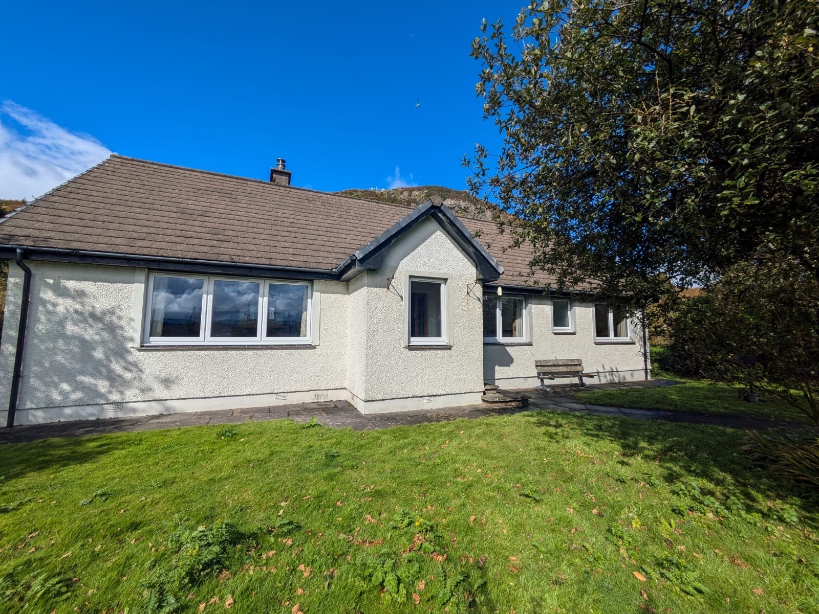 A single-story white house with a brown shingled roof, multiple windows, and a small front porch with a bench, surrounded by a green lawn and trees under a clear blue sky.