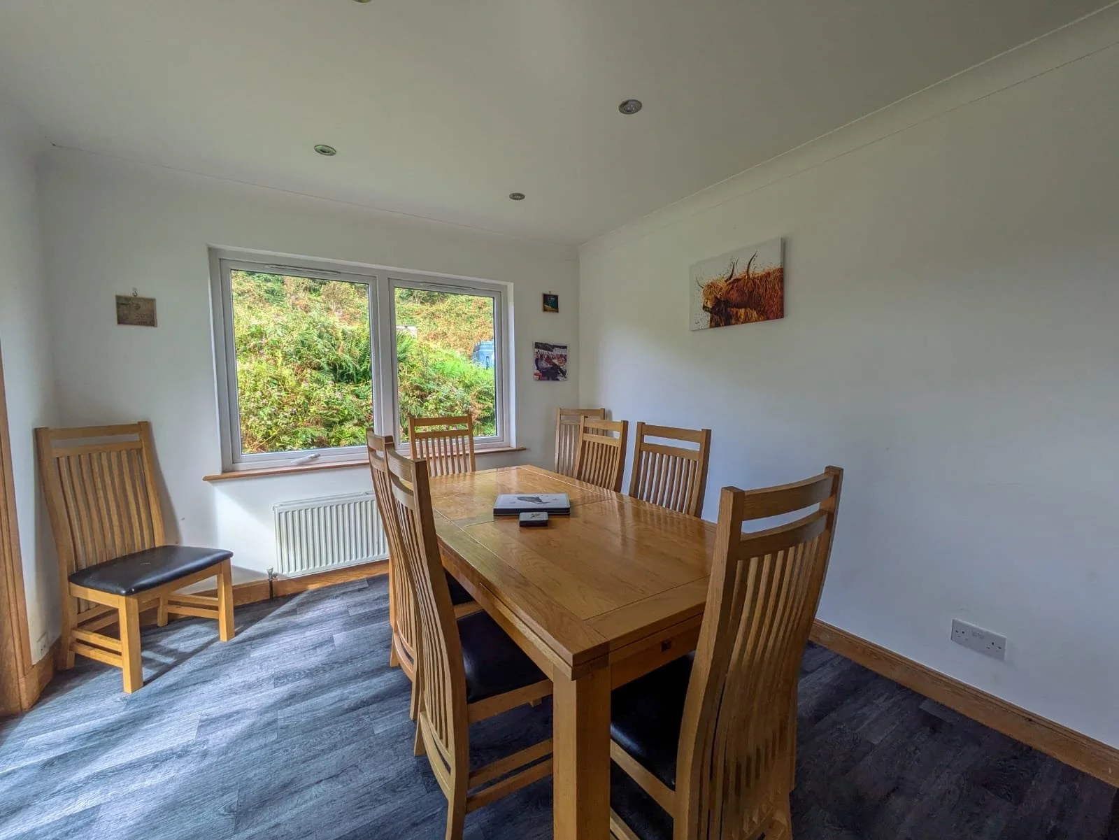 Empty dining room with a wooden table and six matching chairs, window overlooking greenery, paintings on white walls, dark wood flooring.