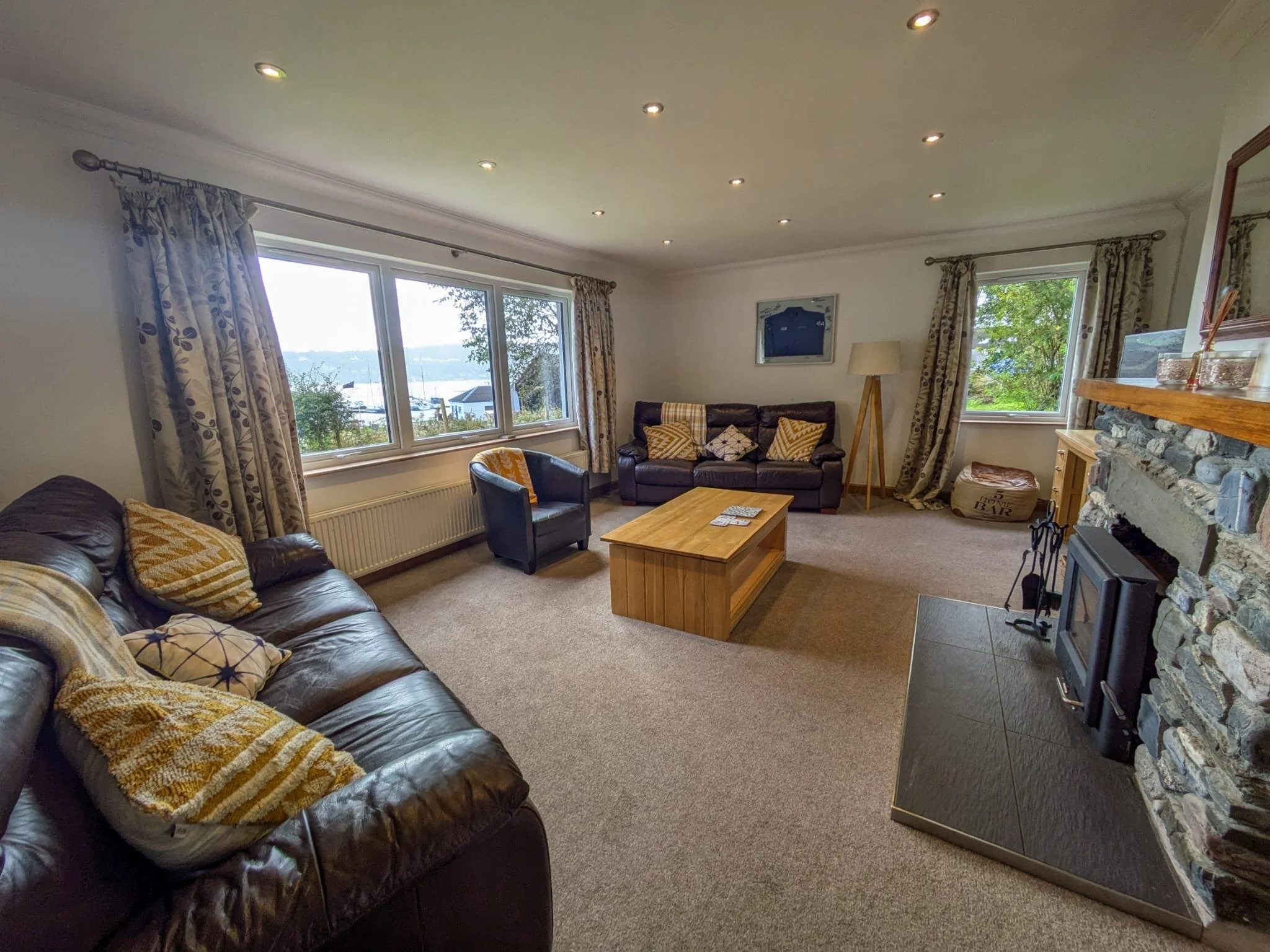 Living room with large front window showing water view, stone fireplace, leather couches, a coffee table, and curtains.