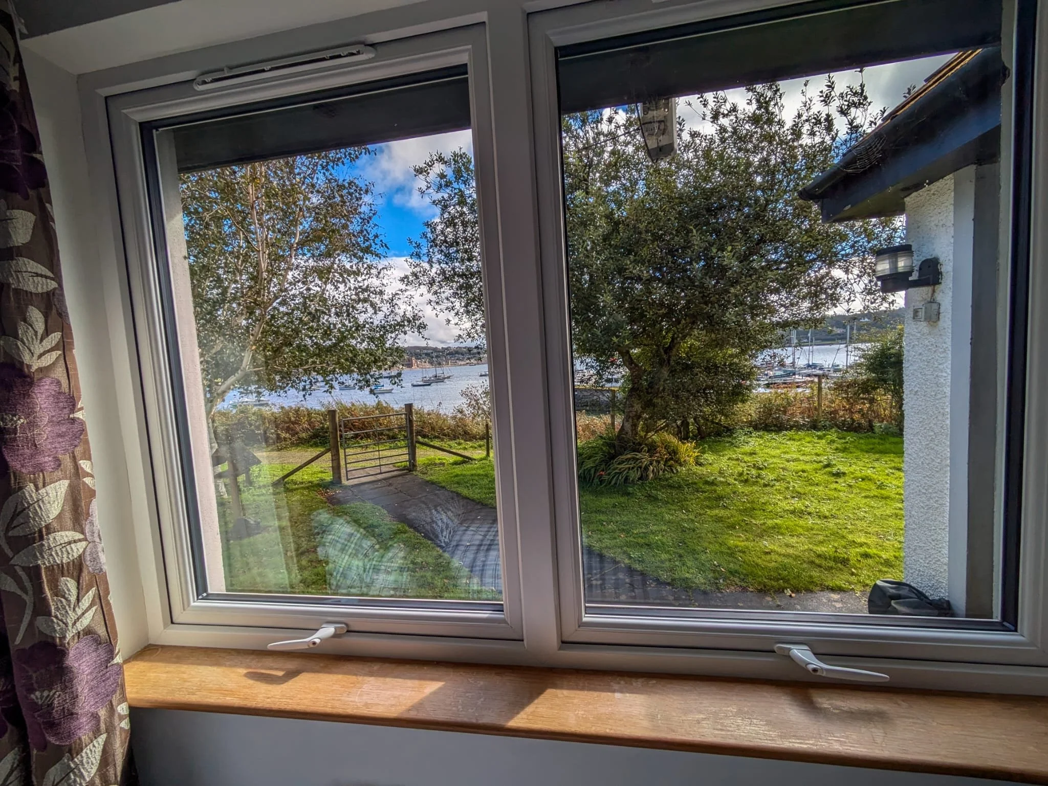 View through a window showing a grassy yard with trees and a body of water with sailboats in the distance, with a partly cloudy sky.