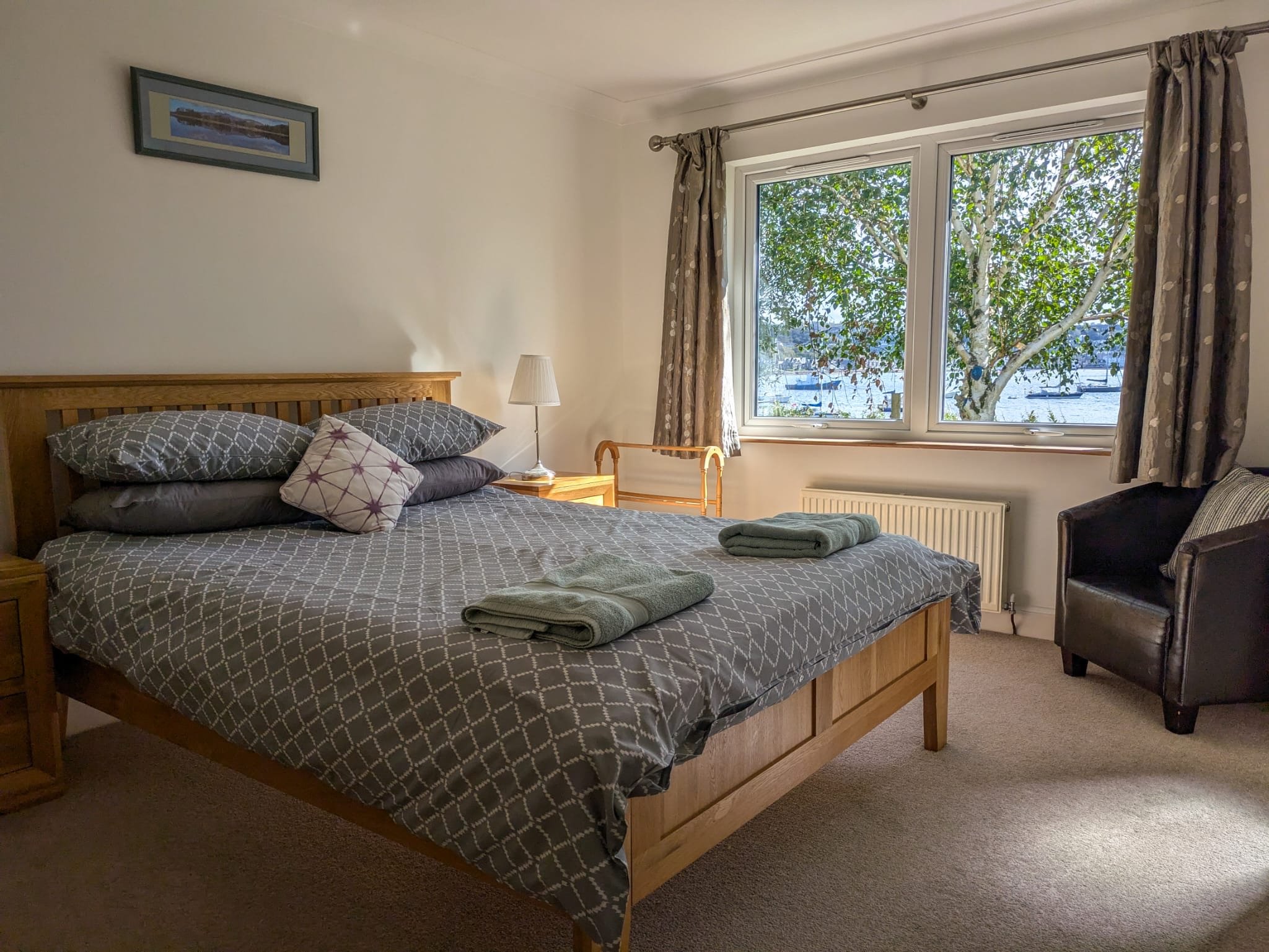 A bedroom with a large wooden bed covered in a patterned gray bedspread and several pillows. A nightstand holding a lamp and a window showing a view of trees and water outside. There is a black armchair next to the window, curtains, and folded towels