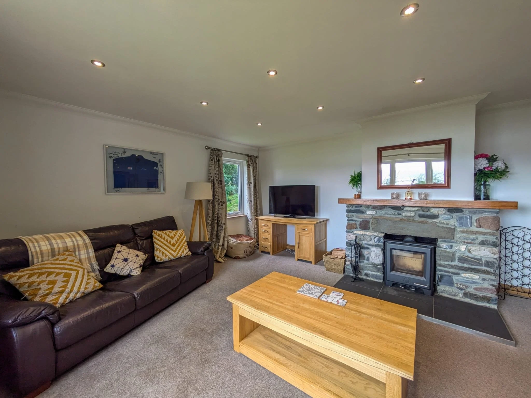 Living room with brown leather sofa, wooden coffee table, stone fireplace with wooden mantel, TV on wooden stand, window with curtains, potted plants, and chandelier.