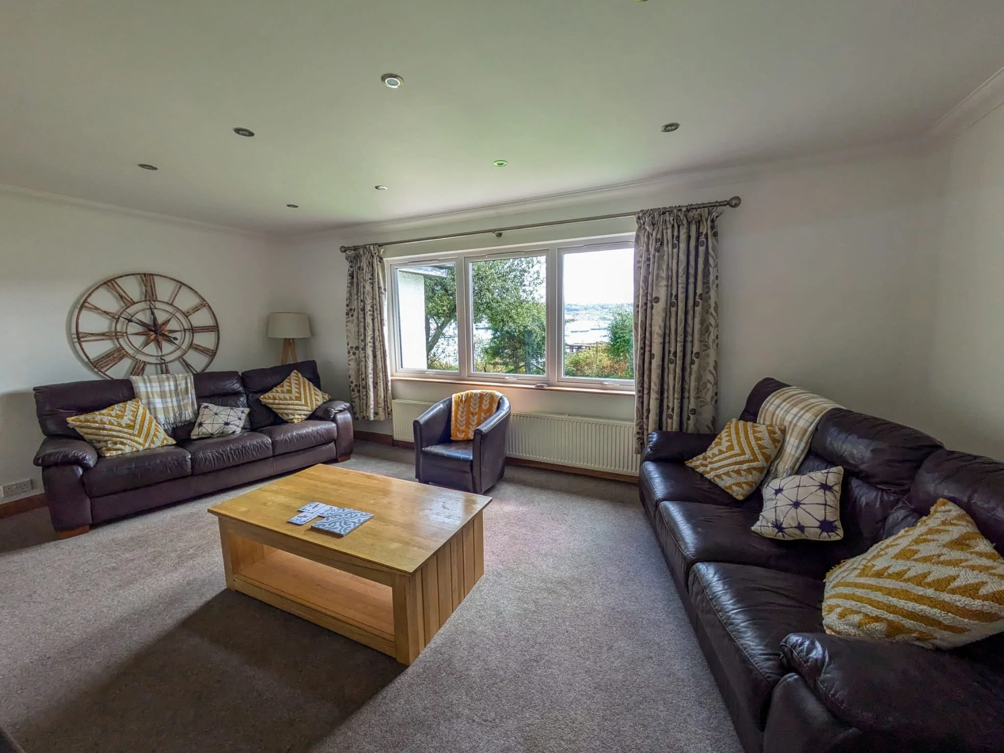 Living room with two dark leather sofas, yellow and white patterned pillows, a wooden coffee table, a large wall clock, a lamp, and a view of trees outside through a large window with patterned curtains.