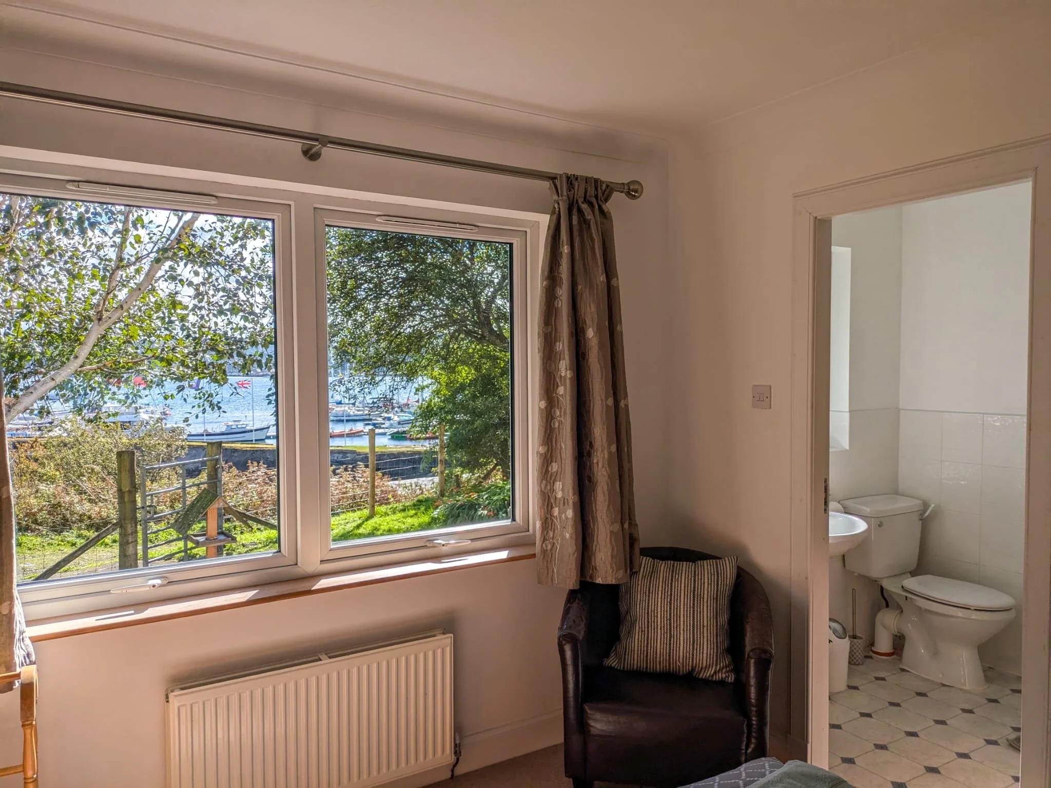 Interior view of a room with a large window showing trees, a fence, and boats on water outside, a small armchair with a striped pillow, and a door leading to a bathroom with a toilet.