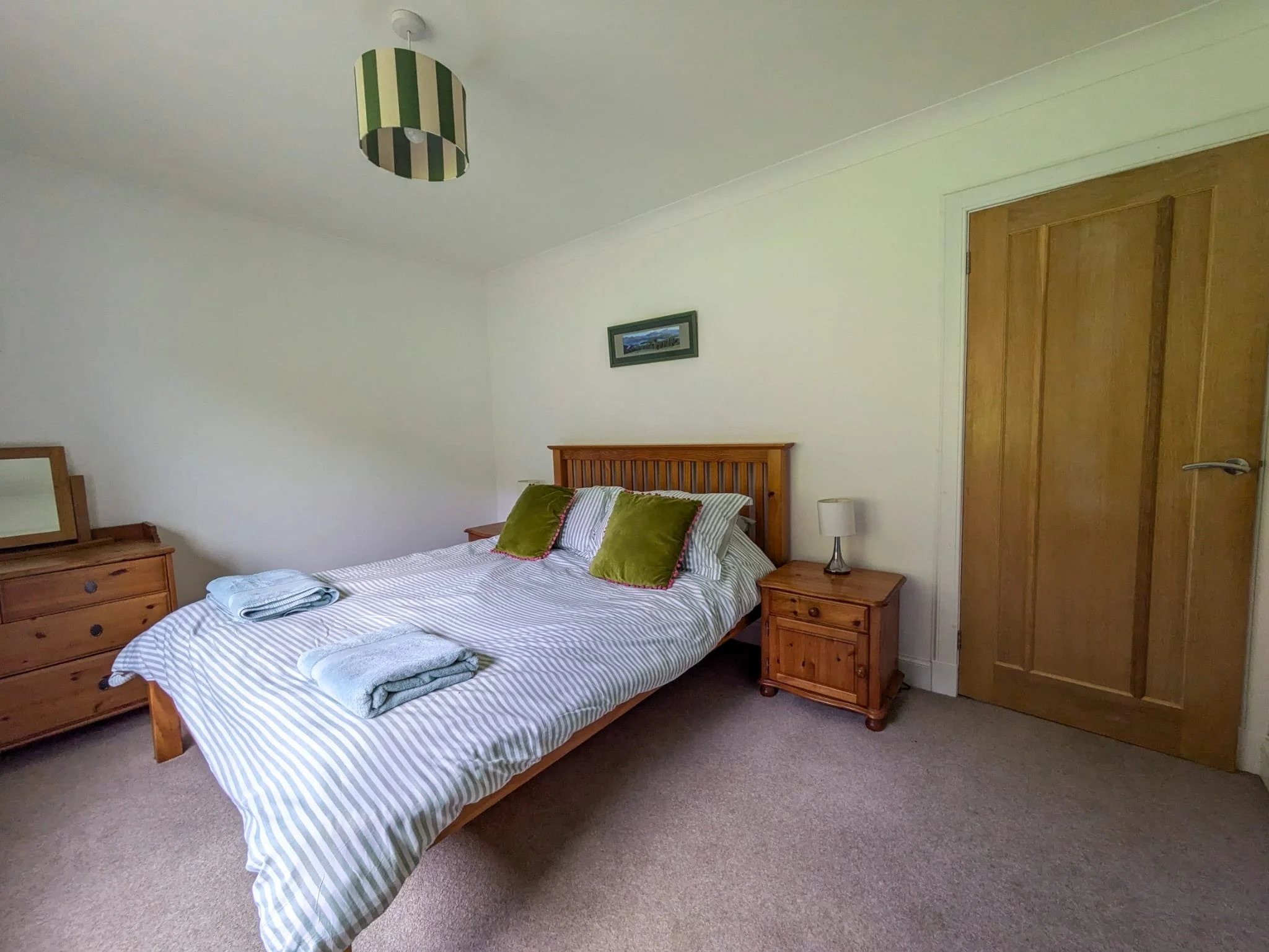 A bedroom with a wooden bed frame and two green pillows on a striped bedspread. There are two folded towels on the bed. To the right of the bed is a wooden nightstand with a small lamp, and to the left is a matching dresser with a mirror. The room ha