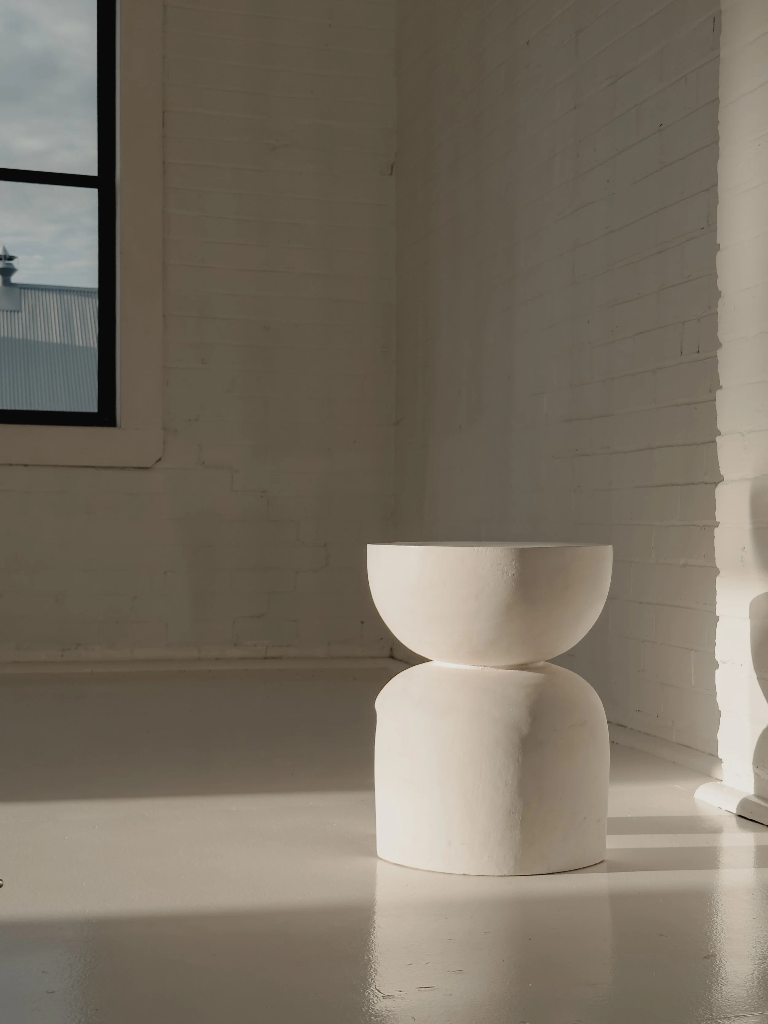 A minimalist white sculptural stool or side table in a bright room with white brick walls and a window showing a cloudy sky.