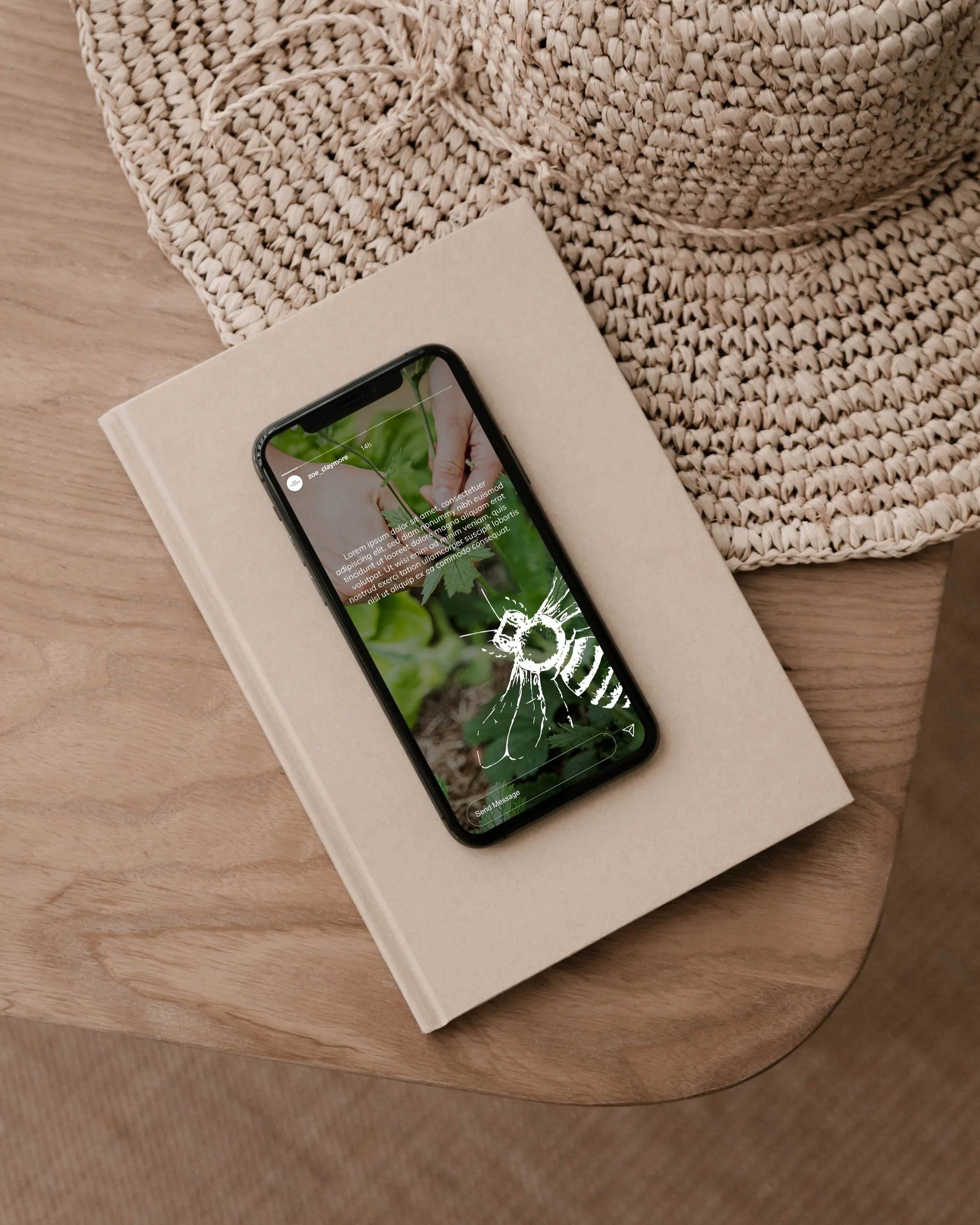 Smartphone displaying a bee on a green leaf with overlaid text, resting on a closed beige book on a wooden table next to a woven hat and a textured fabric item.