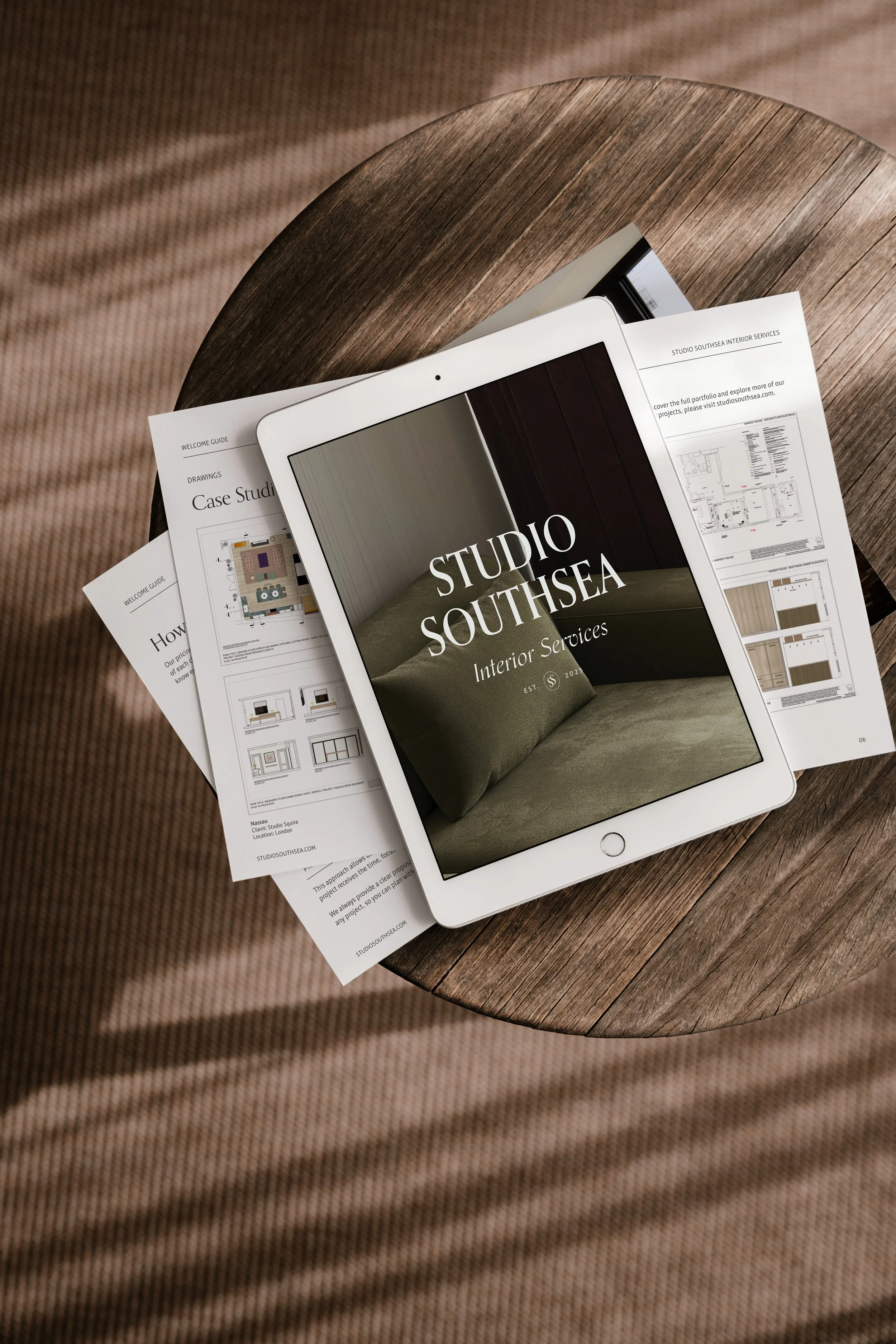 An iPad displaying a cover page for Studio Southsea Interior Services, placed on top of printed interior design plans and brochures on a round wooden table.