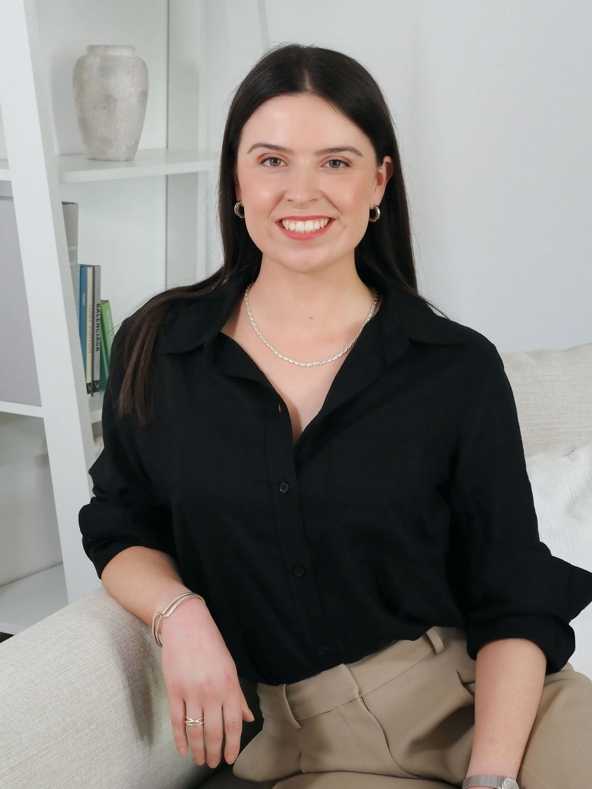 A woman with long dark hair, wearing a black button-up shirt, beige pants, and silver jewelry, sitting on a white couch with a bookshelf and decorative items in the background.