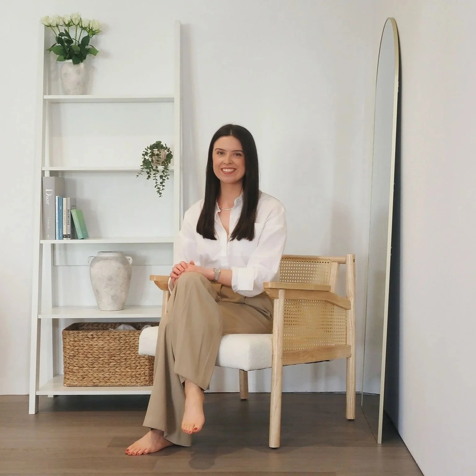 A woman with dark hair smiling, sitting barefoot on a wooden and wicker chair in a minimalist room with white walls, a white bookshelf with decorative plants and books, and a floor-length mirror.