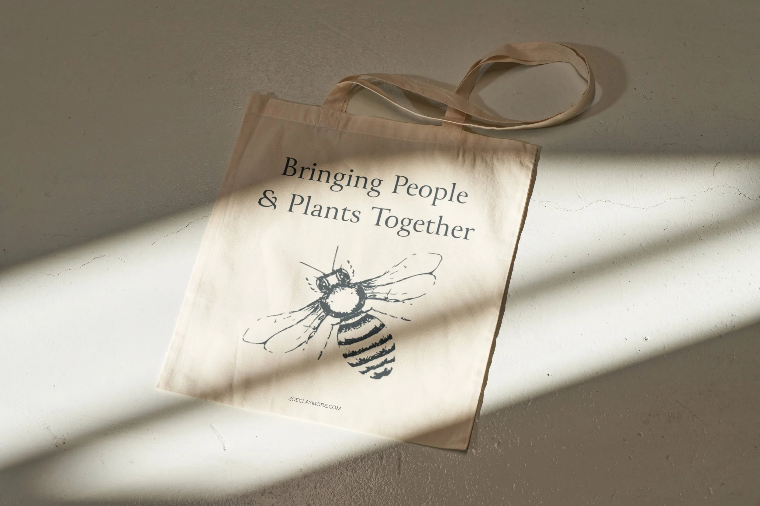 Tan tote bag with the words 'Bringing People & Plants Together' and a bee sketch, on a textured white surface with sunlight and shadow.