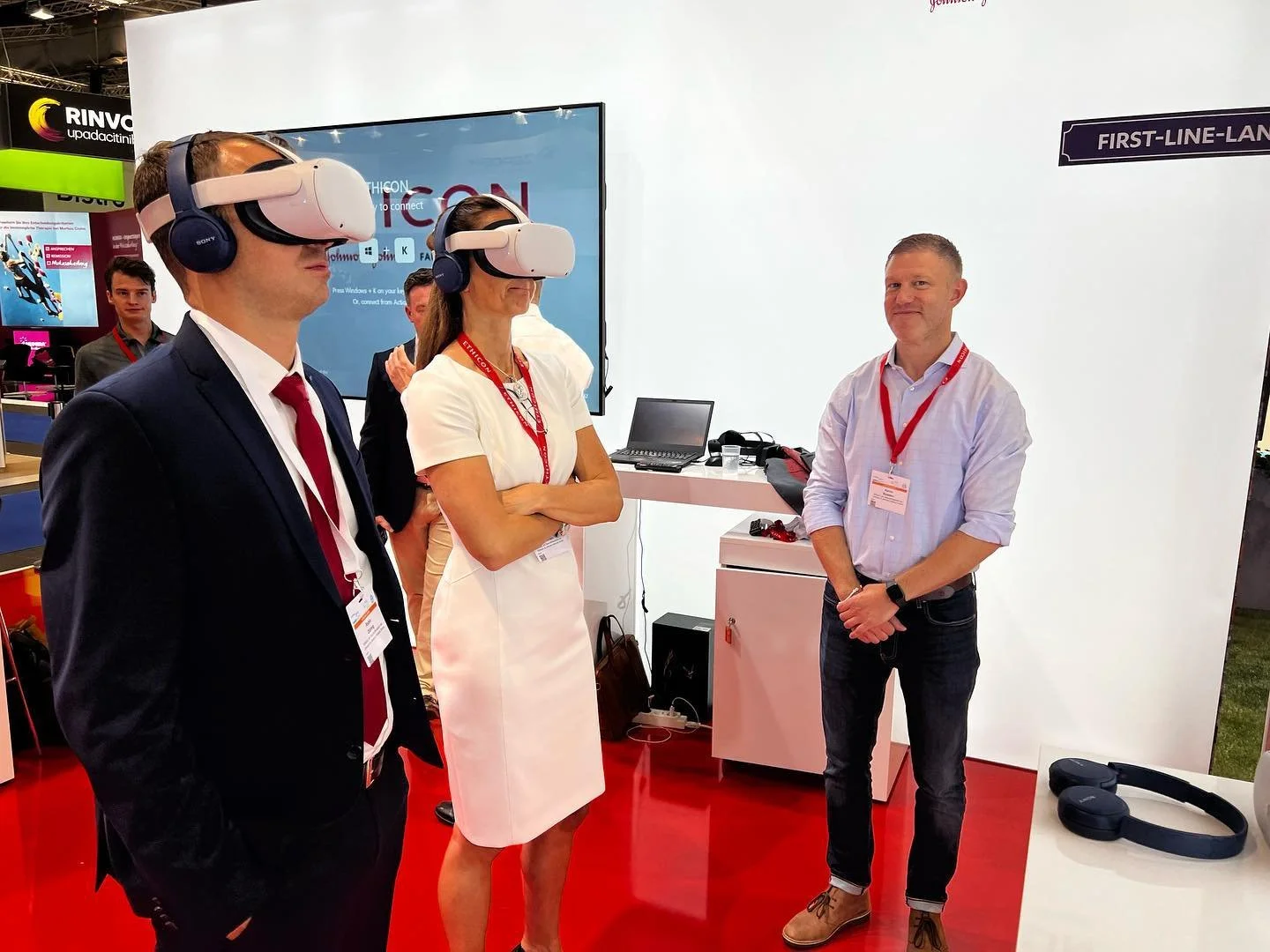 Several people at a trade show, with two individuals wearing virtual reality headsets, standing on a red carpet.