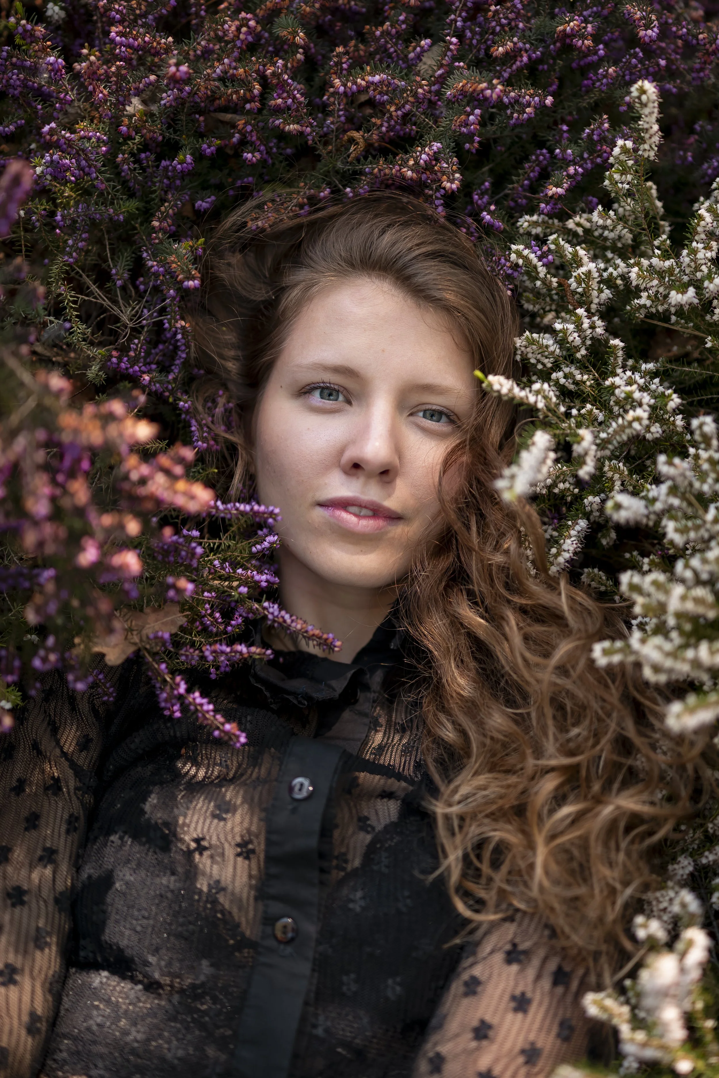 Junge Frau mit lockigem braunem Haar liegt zwischen violetten und weißen Blumen und schaut in die Kamera.