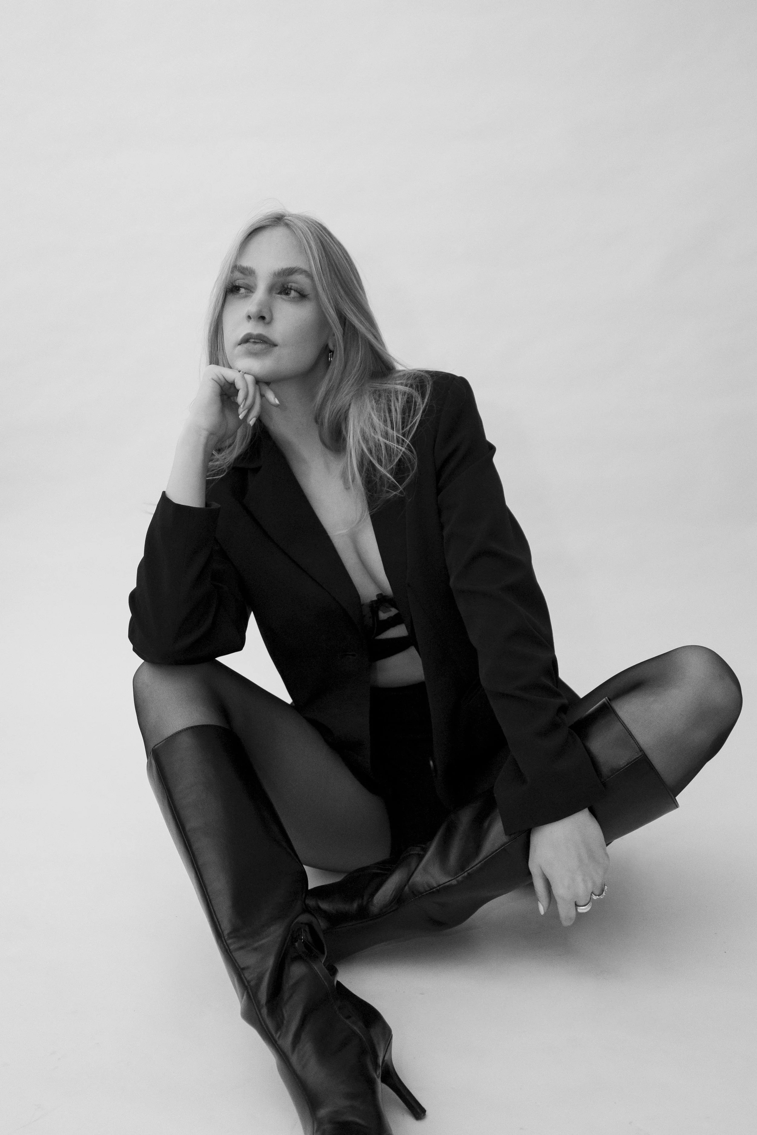 Schwarz-Weiß-Foto einer jungen Frau, die in einem schwarzen Anzug sitzt mit hochhackigen Stiefeln, entspannt und nachdenklich wirkend.