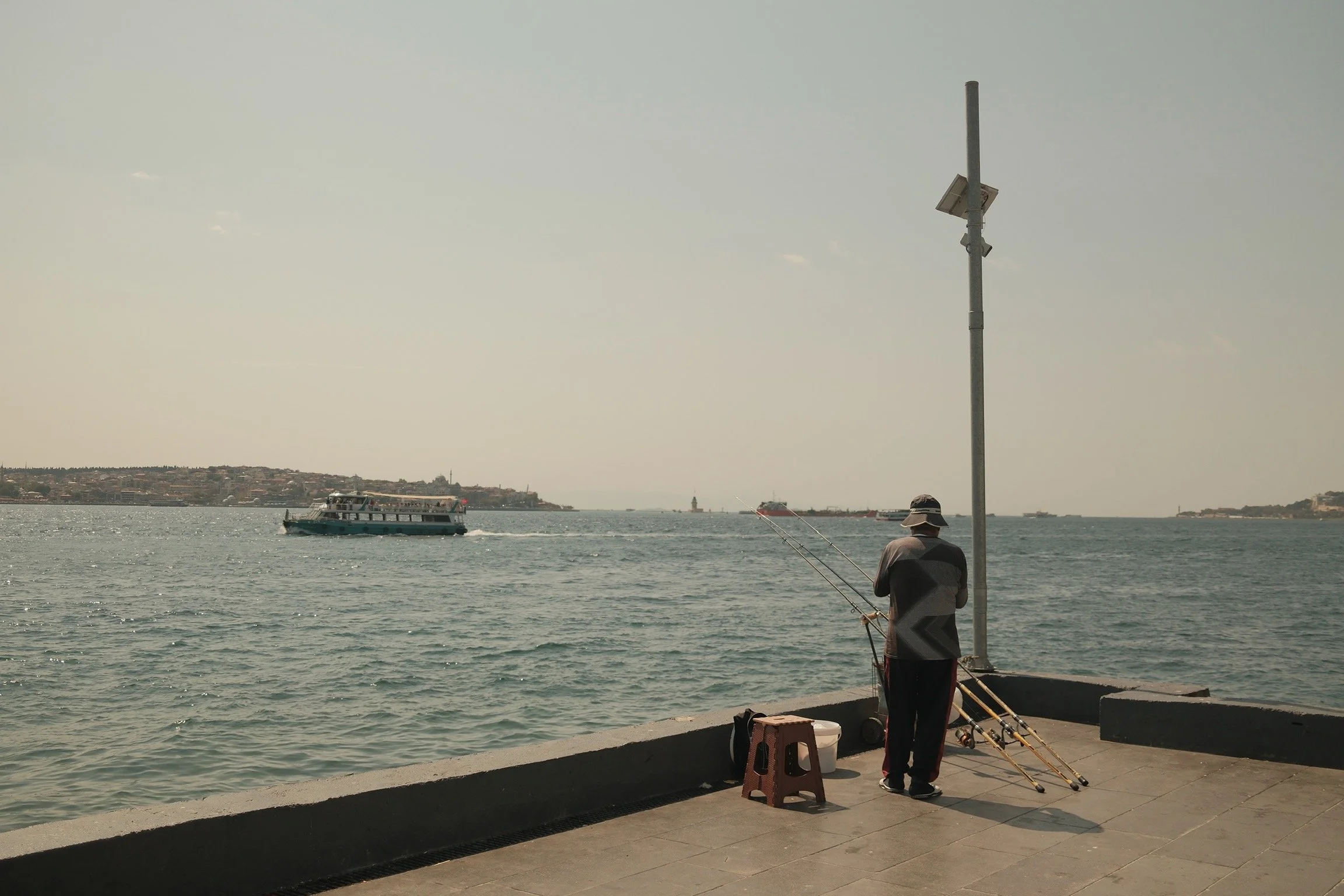Ein Mann steht mit Angelausrüstung an einer Hafenpromenade, blickt aufs Wasser, während Boote vorbeifahren. Es ist sonnig, und der Himmel ist leicht bewölkt.