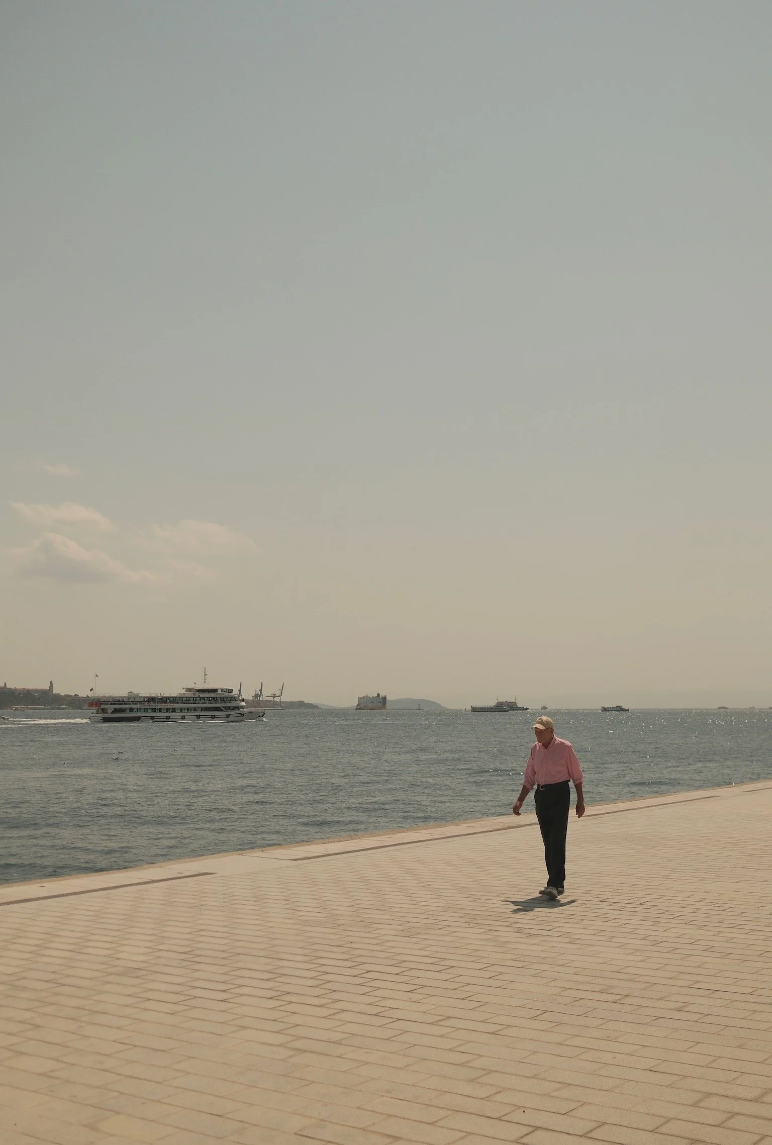 Eine Person, die am Wasser entlang spaziert, bei sonnigem Himmel. Im Wasser fahren Schiffe und Boote.