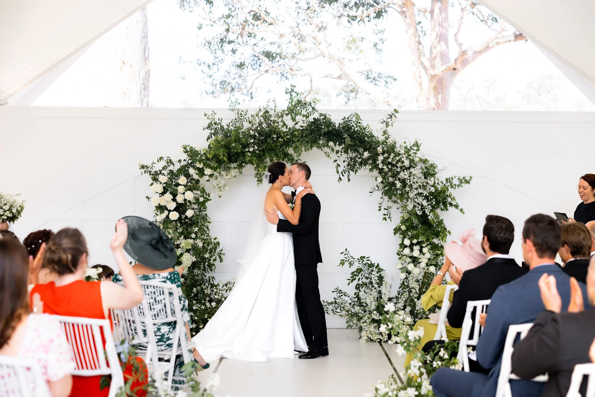 Two women in wedding dresses sharing a joyful moment among lush green foliage, holding a bouquet of flowers.
