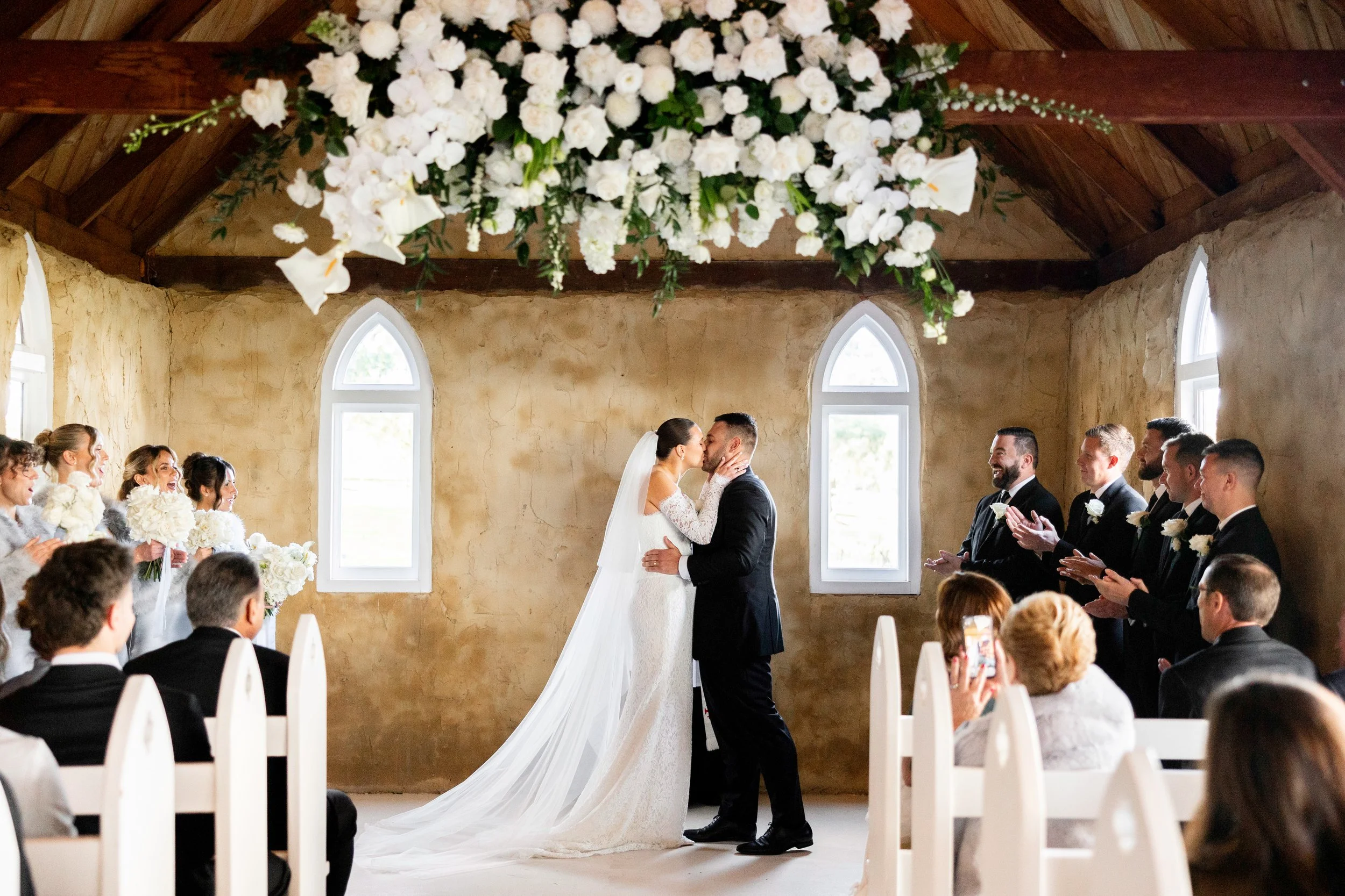 A bride and groom standing in a decorated wedding reception hall with a large floral arrangement and a chandelier hanging from a wooden ceiling.