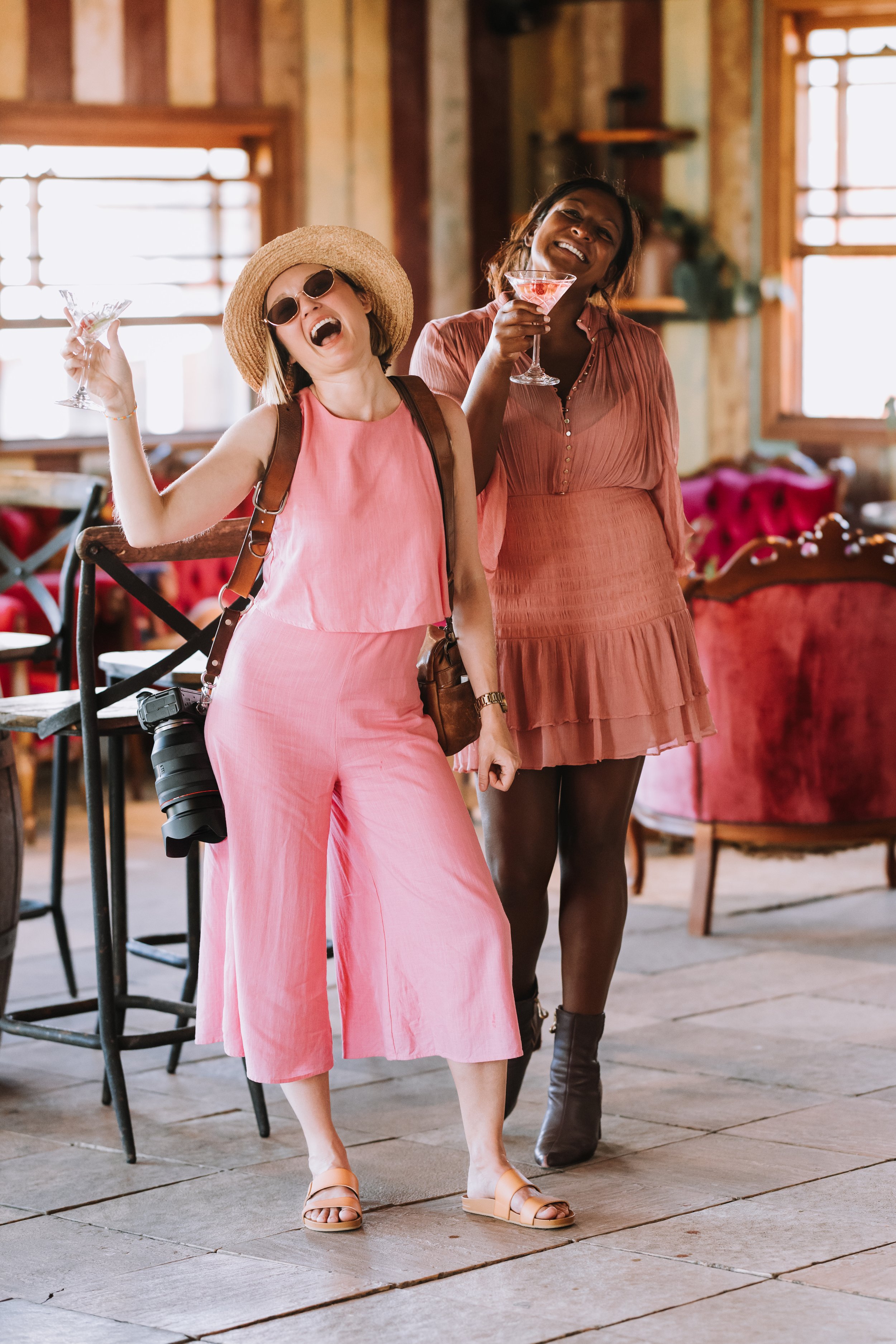 Two women enjoying drinks and dancing in a rustic, cozy restaurant with wooden decor and large windows.